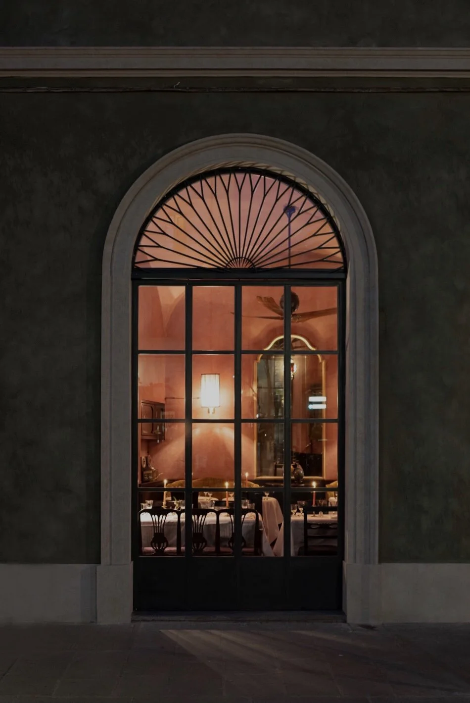 View through a large glass door revealing a warmly lit dining room with a long table, elegant chairs, a large mirror, and a chandelier, with pink walls and decorative elements.