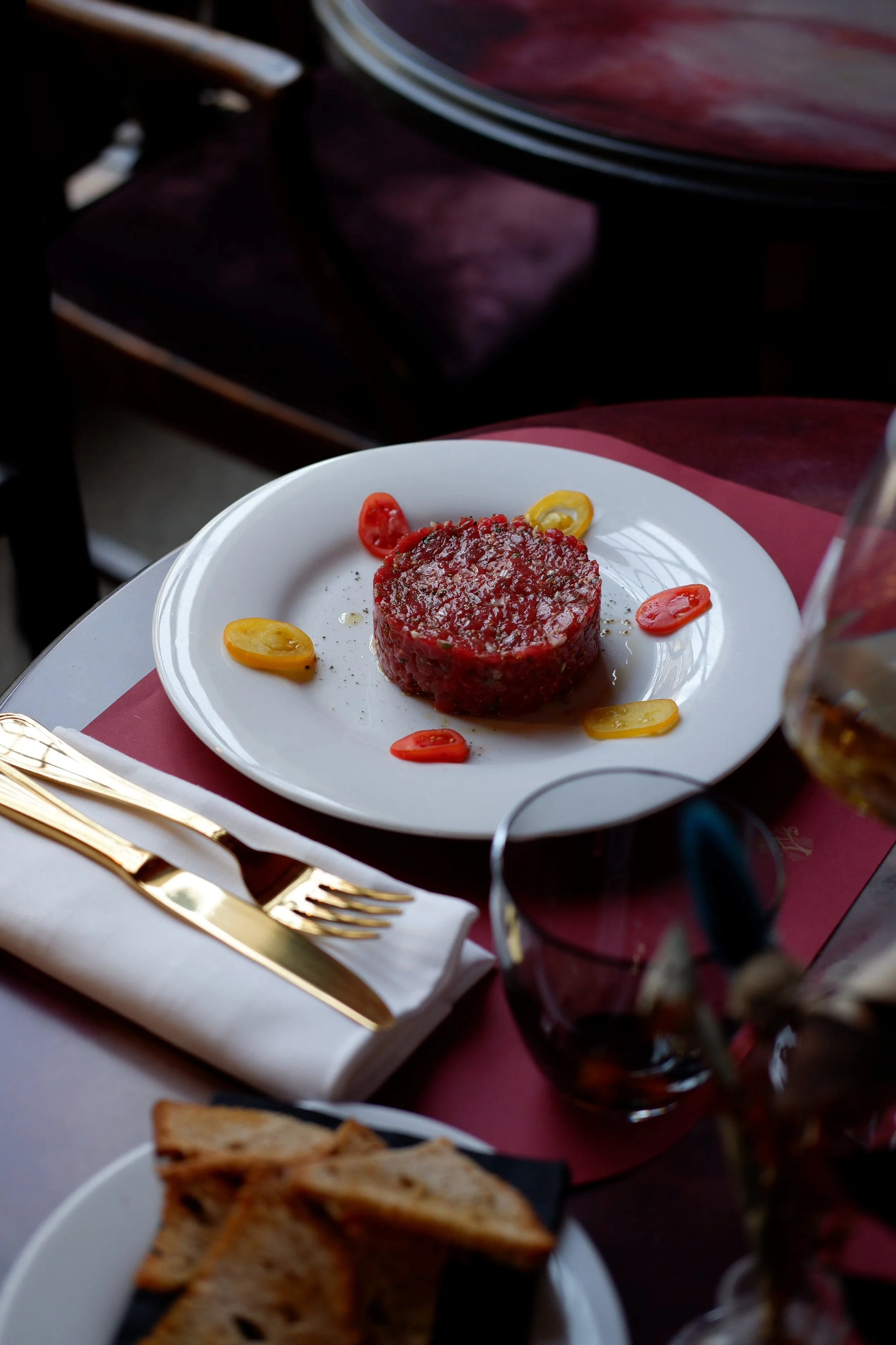 A white plate with a serving of steak tartare topped with black pepper, surrounded by sliced cherry tomatoes in red and yellow, on a table with gold cutlery and a white napkin, at a restaurant.