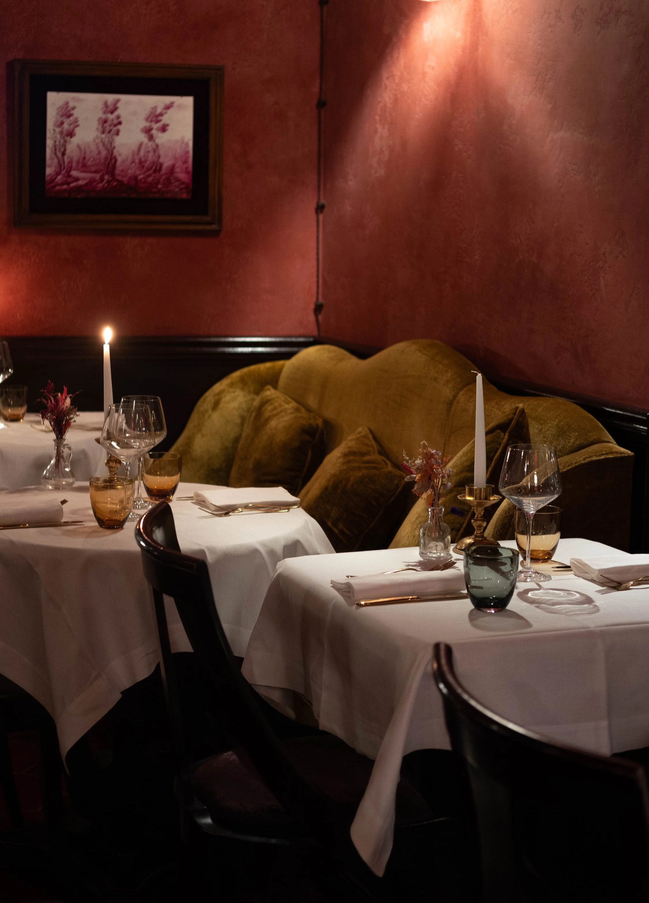 Elegant restaurant table setting with white tablecloths, candles, glassware, and place settings, against a red textured wall with framed artwork and a yellow velvet sofa.