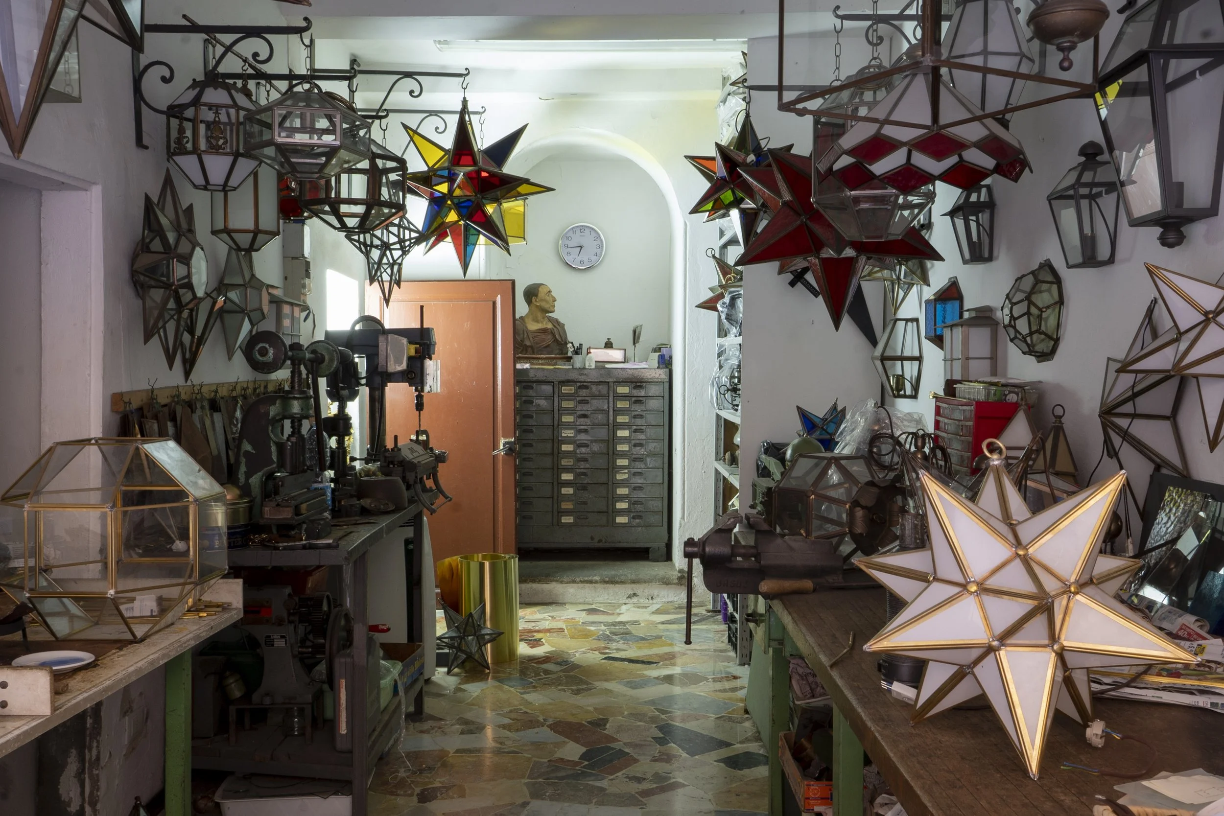 Workshop with hanging decorative lamps and star-shaped lanterns, cluttered workbenches, tools, storage drawers, and a bust sculpture on a cabinet at the back.