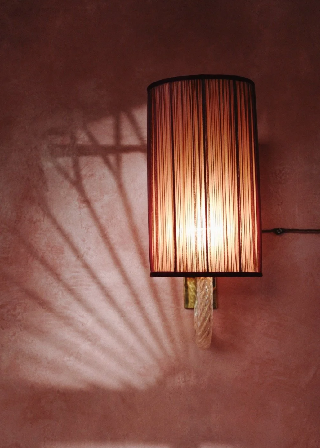 A wall-mounted cylindrical lampshade casting a patterned shadow on a pink wall.