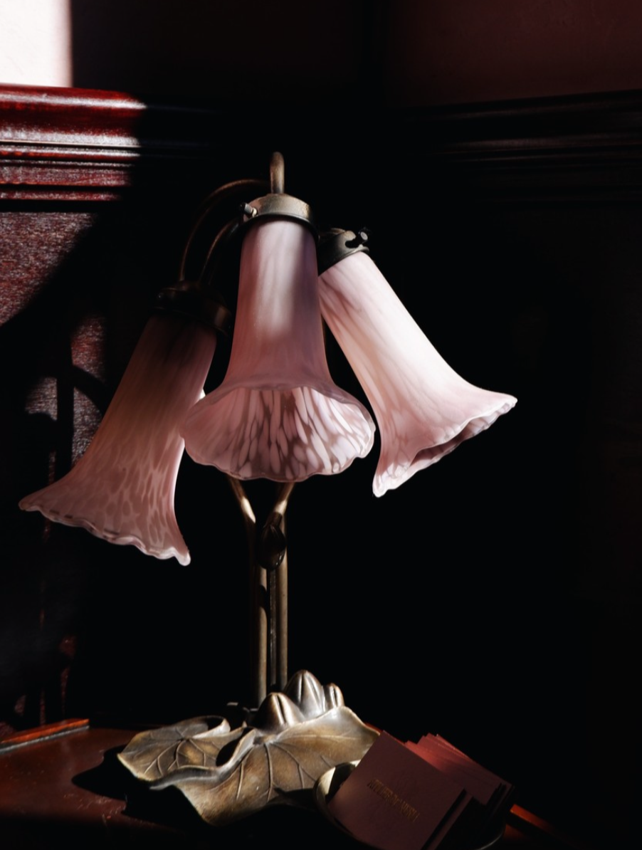 An ornate table lamp with three pink floral-shaped glass shades, gold-colored metal base, and leaf-shaped decorative elements on a dark wooden surface.