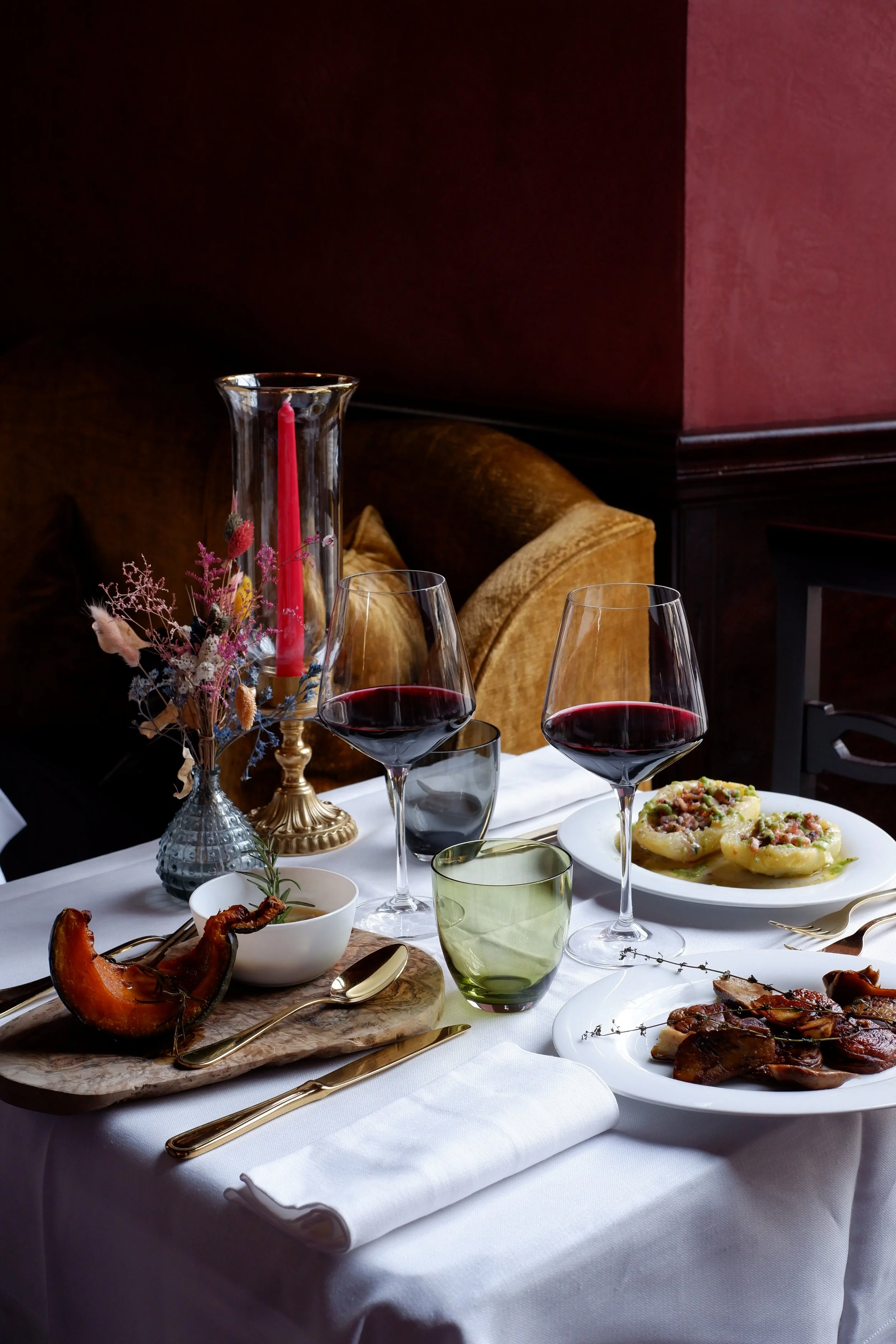 A elegantly set dinner table with wine glasses, a candle, flowers, and various dishes including roasted meat, ravioli, and roasted vegetables.
