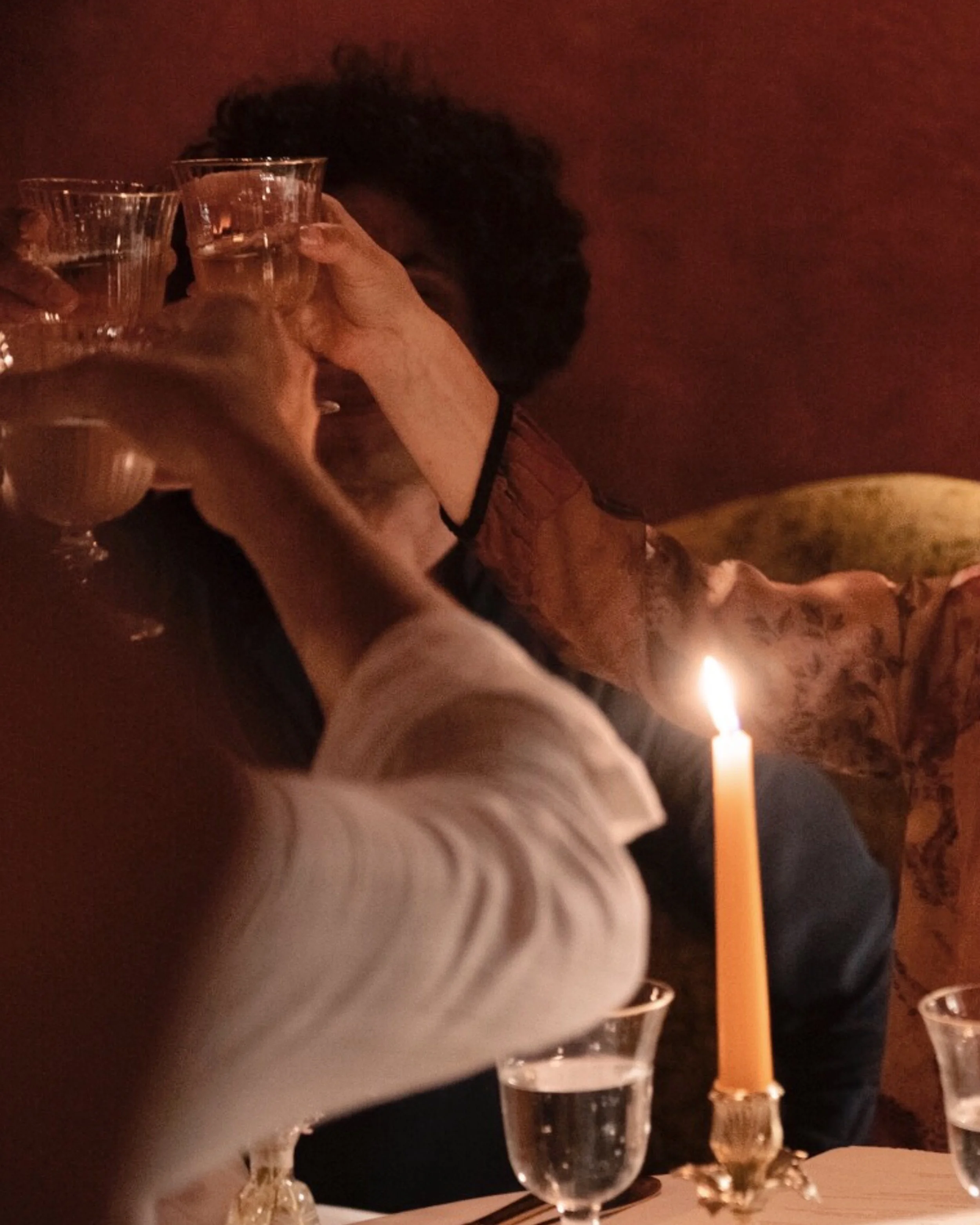 People celebrating around a table with wine glasses and a lit candle in a dimly lit setting, possibly a restaurant or gathering.