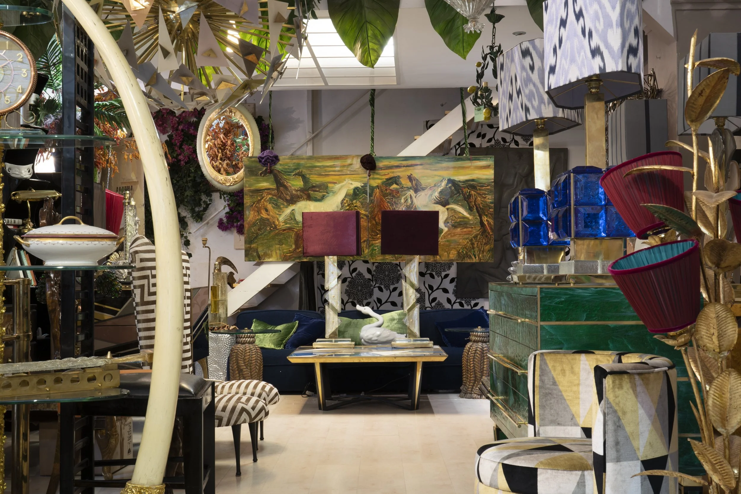 Eclectic interior decor with various lamps, sculptures, and colorful furniture, featuring a large painting of horses and swans in the background.