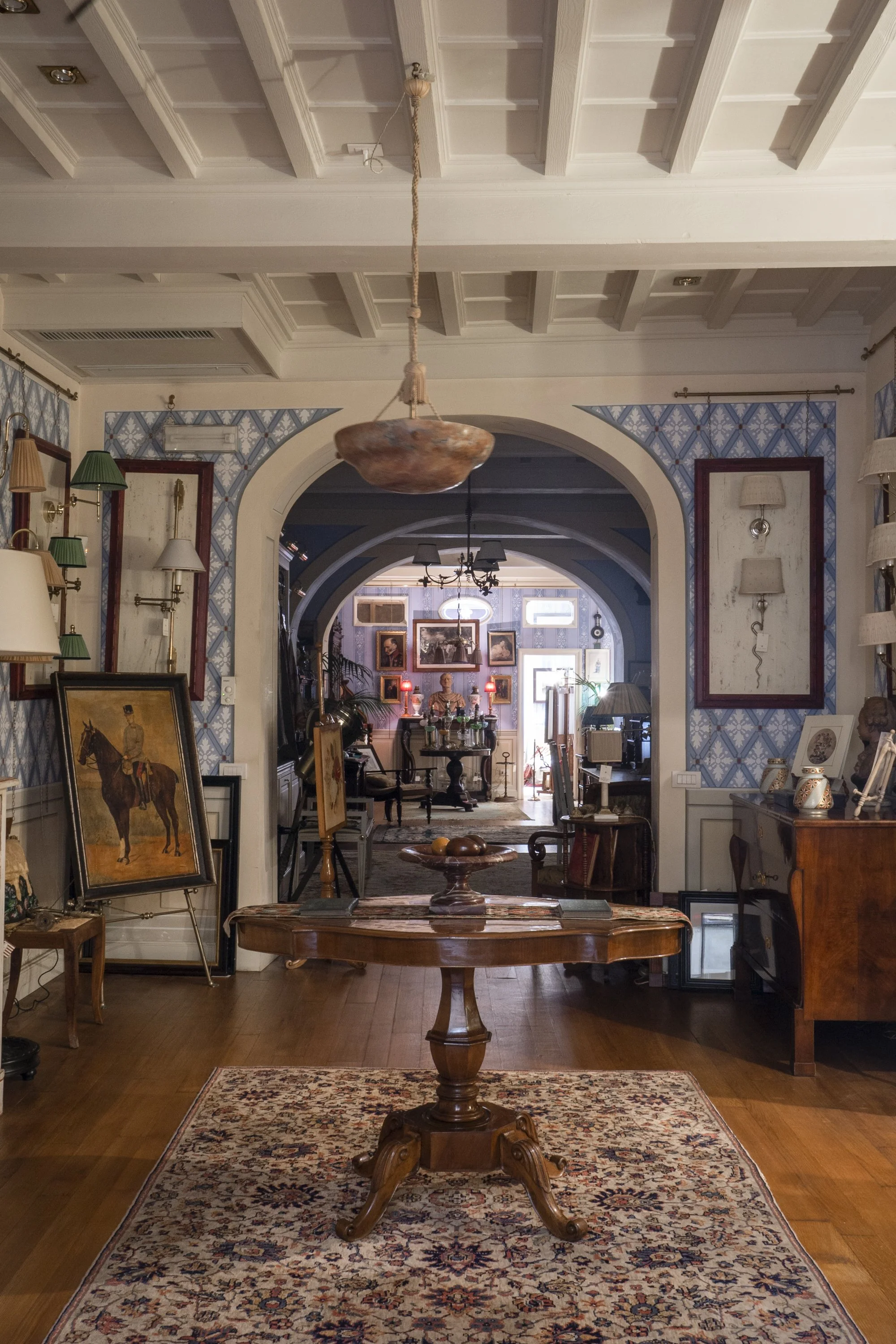 An antique-style wooden table in the center of a room with a patterned rug beneath it. The room has vintage artwork, framed photographs, lamps, and decorative items along the walls and furniture. Arched doorways lead to another room with more artwork and furnishings.