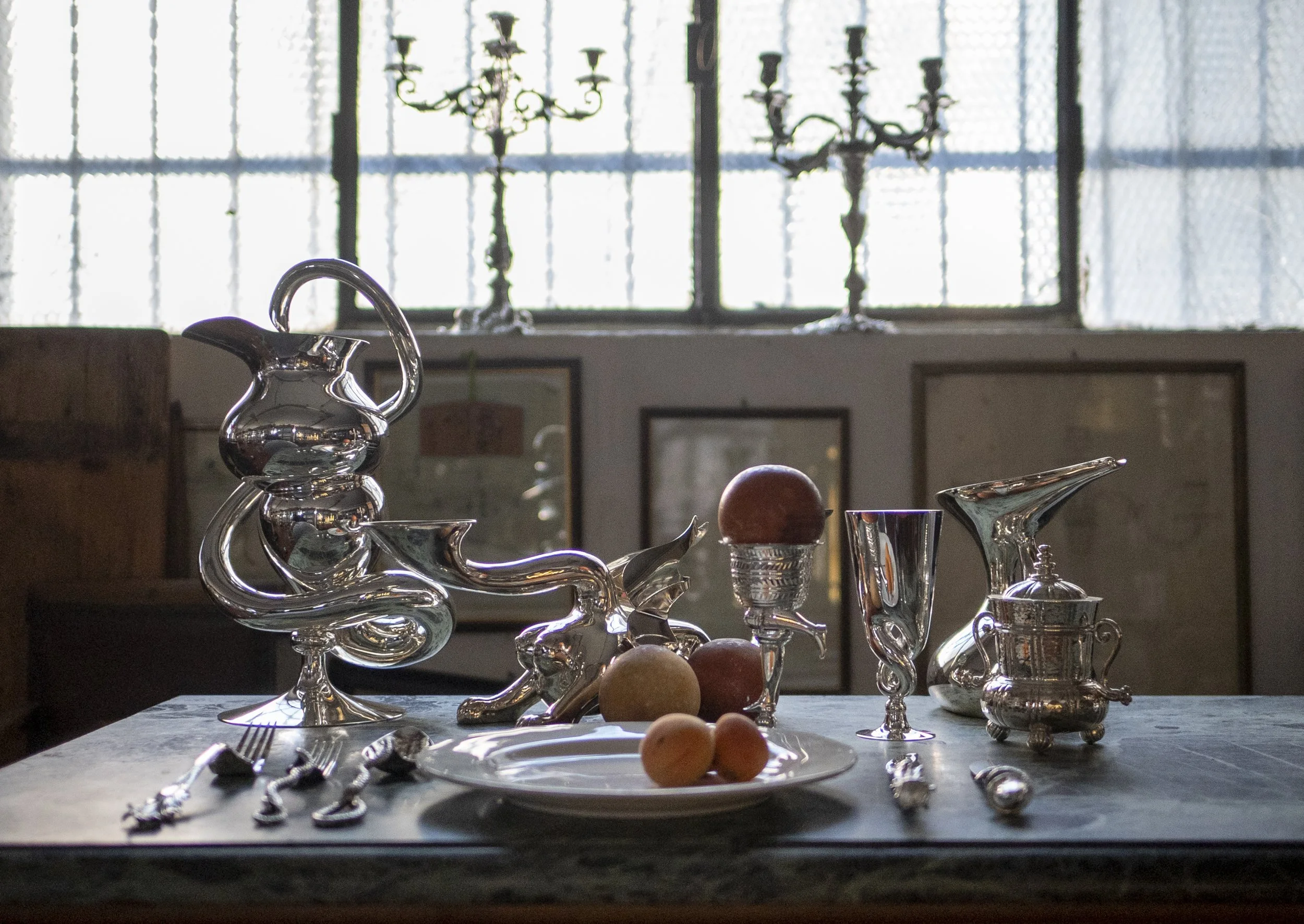 Table with silver decorative items, fruits, and utensils in a room with large windows and candelabras in the background.