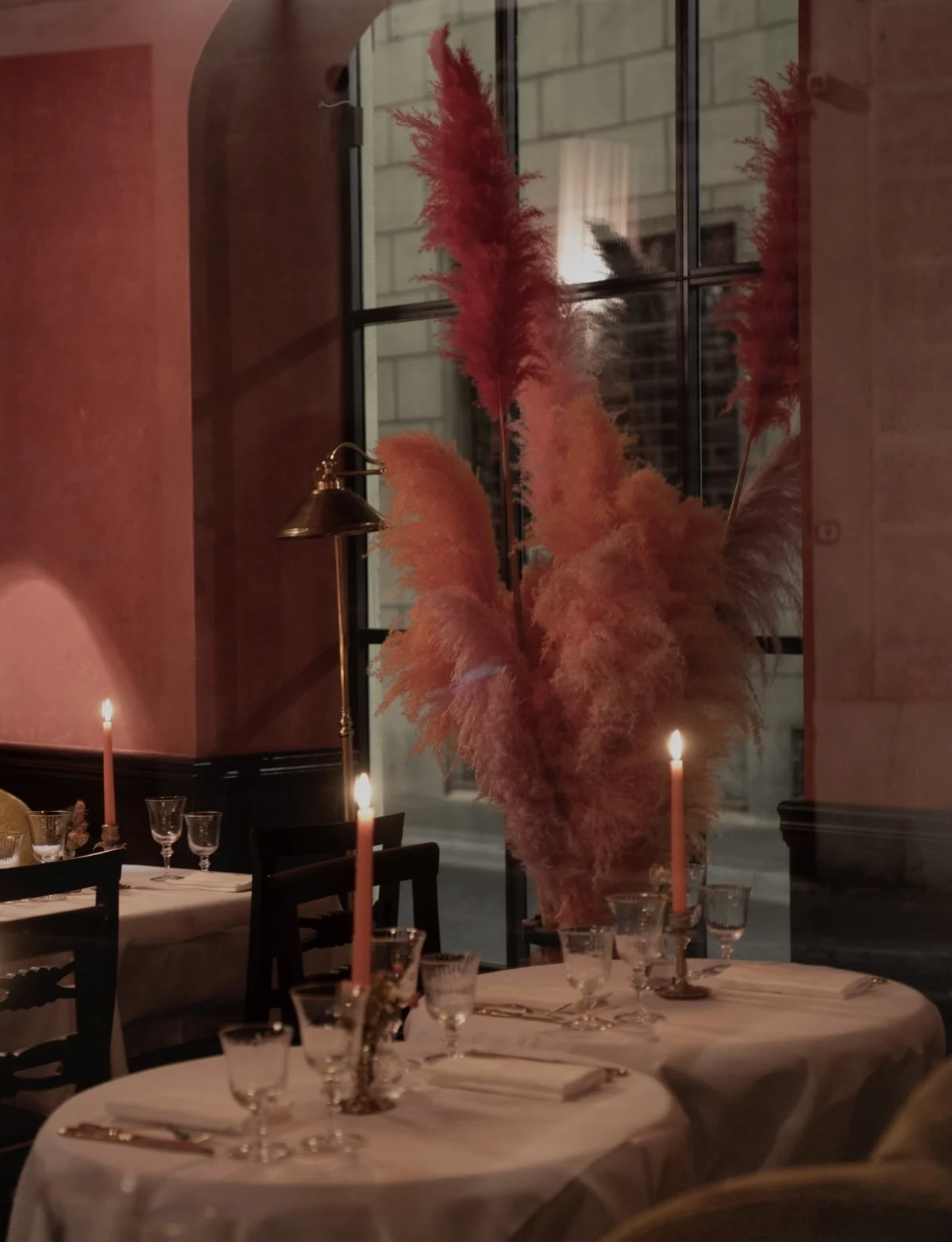 Elegant dining table with a large pink and red feather centerpiece, candlelit candles, wine glasses, and napkins, inside a sophisticated restaurant with large windows showing a cityscape outside.
