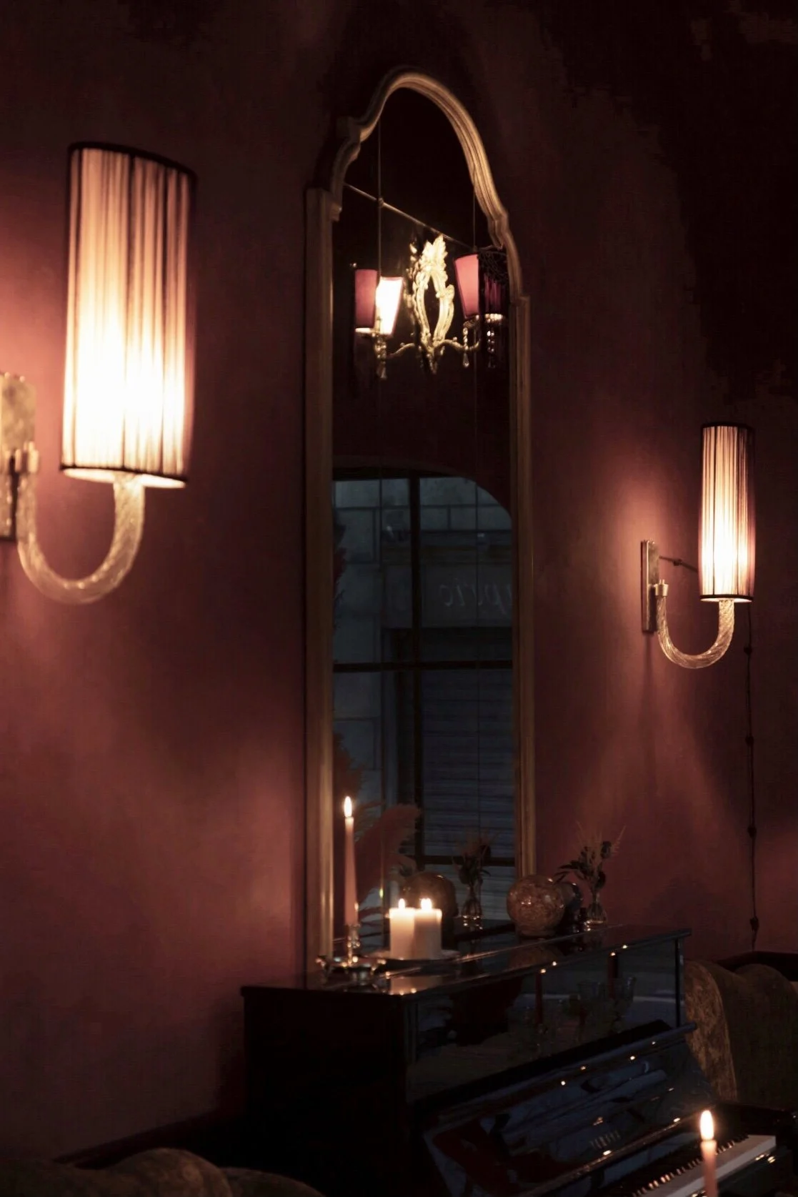Dimly lit room with pinkish walls, two wall sconces with elongated lampshades, a tall mirror with an ornate frame, a piano with lit candles and vases with flowers reflected in the mirror.