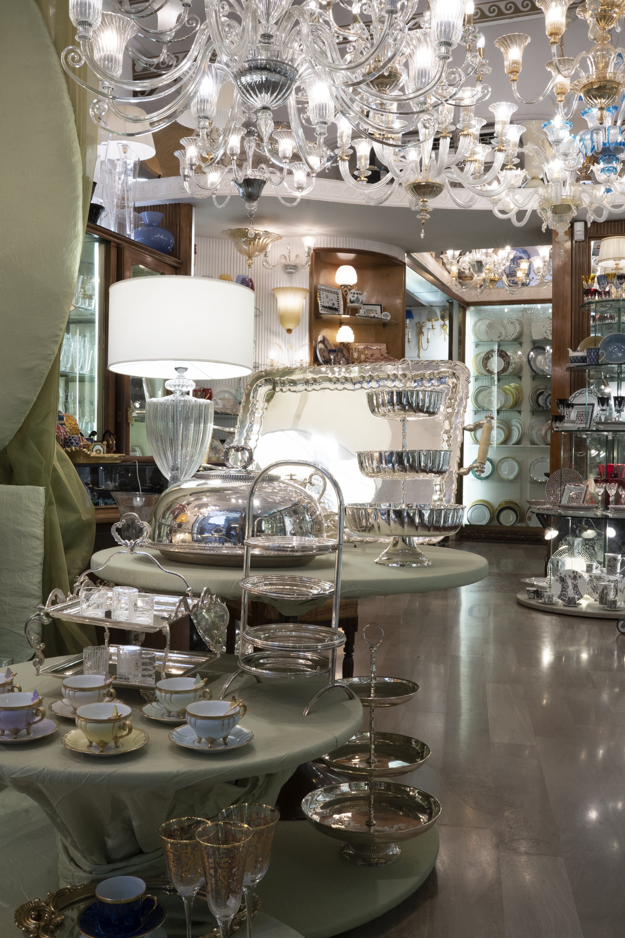 A display of glassware, china, and decorative items in a well-lit antique shop with chandeliers and wooden shelves.