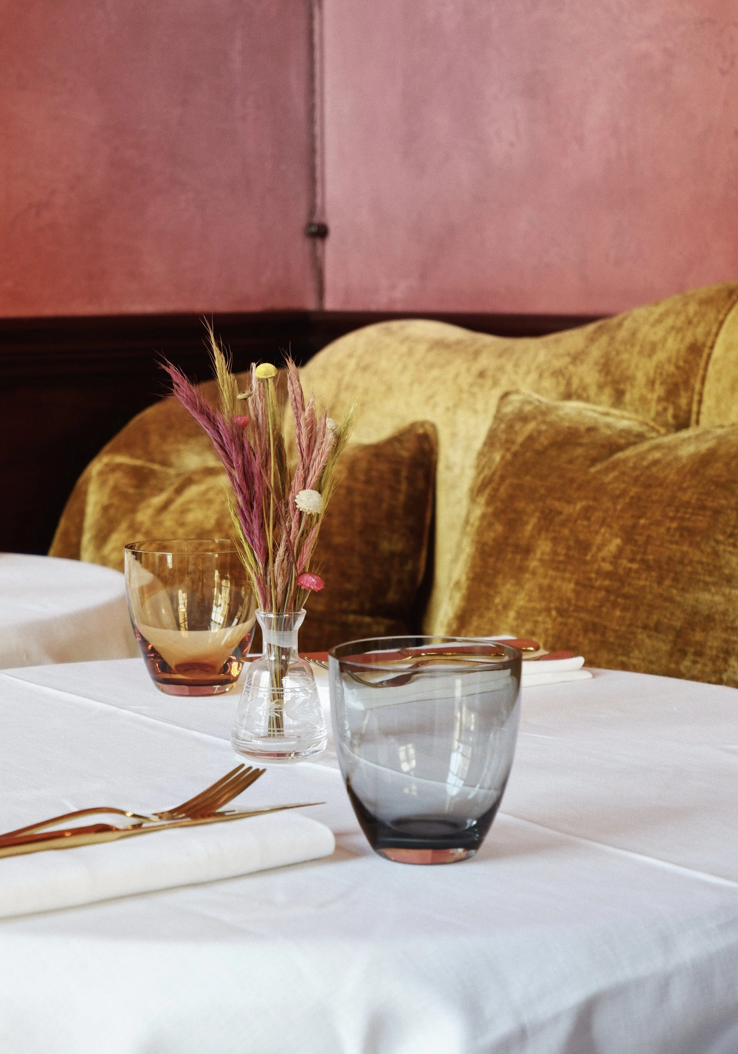 A restaurant table setting with pink, amber, and gray drinking glasses, a small vase with pink and yellow dried flowers, a white napkin with a fork, and a cushioned yellow-brown upholstered bench against a pink wall.