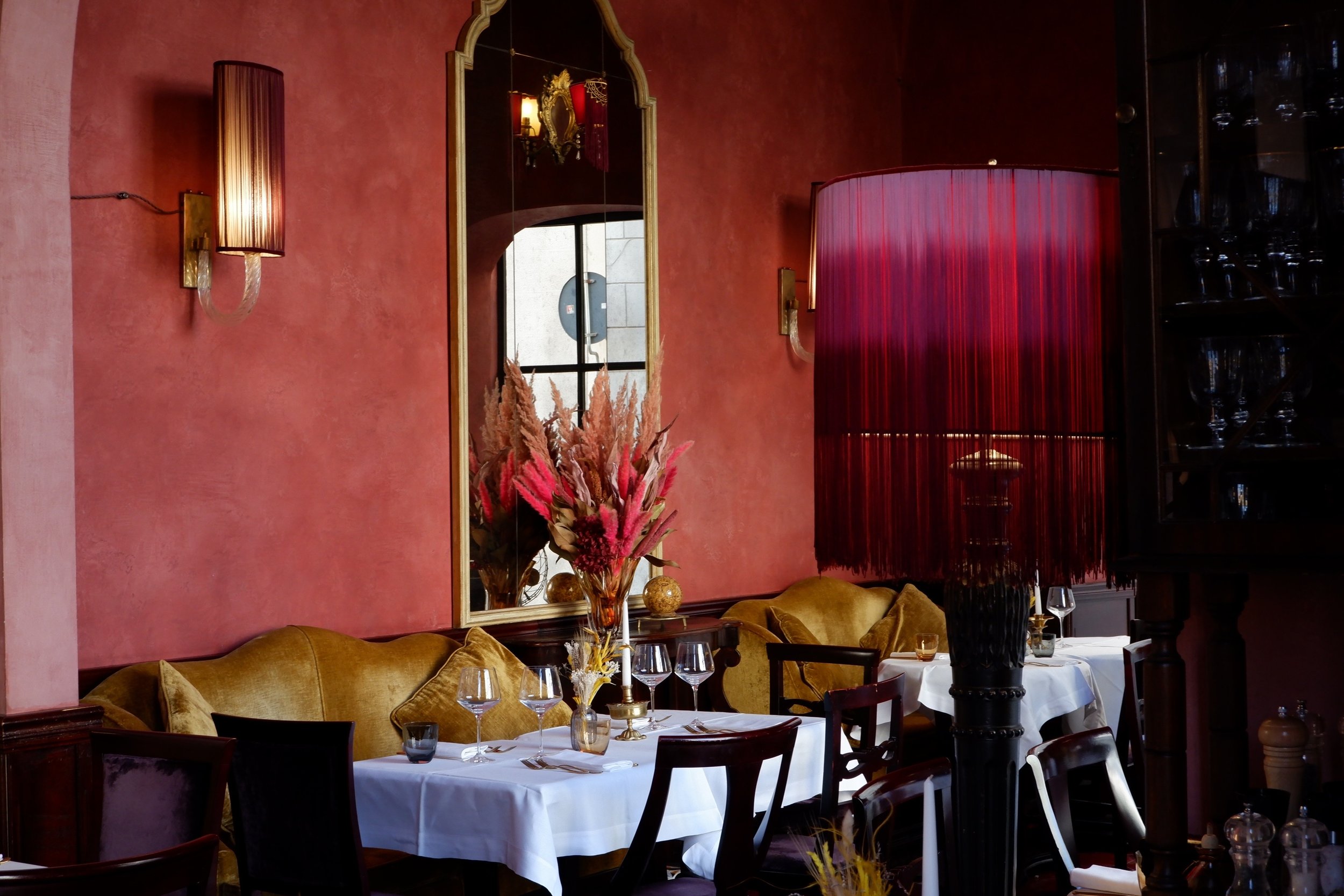Elegant restaurant interior with pink textured walls, gold accents, a large mirror, and a floral centerpiece with pink and beige dried flowers on a dining table dressed with white tablecloths, glassware, and candles.