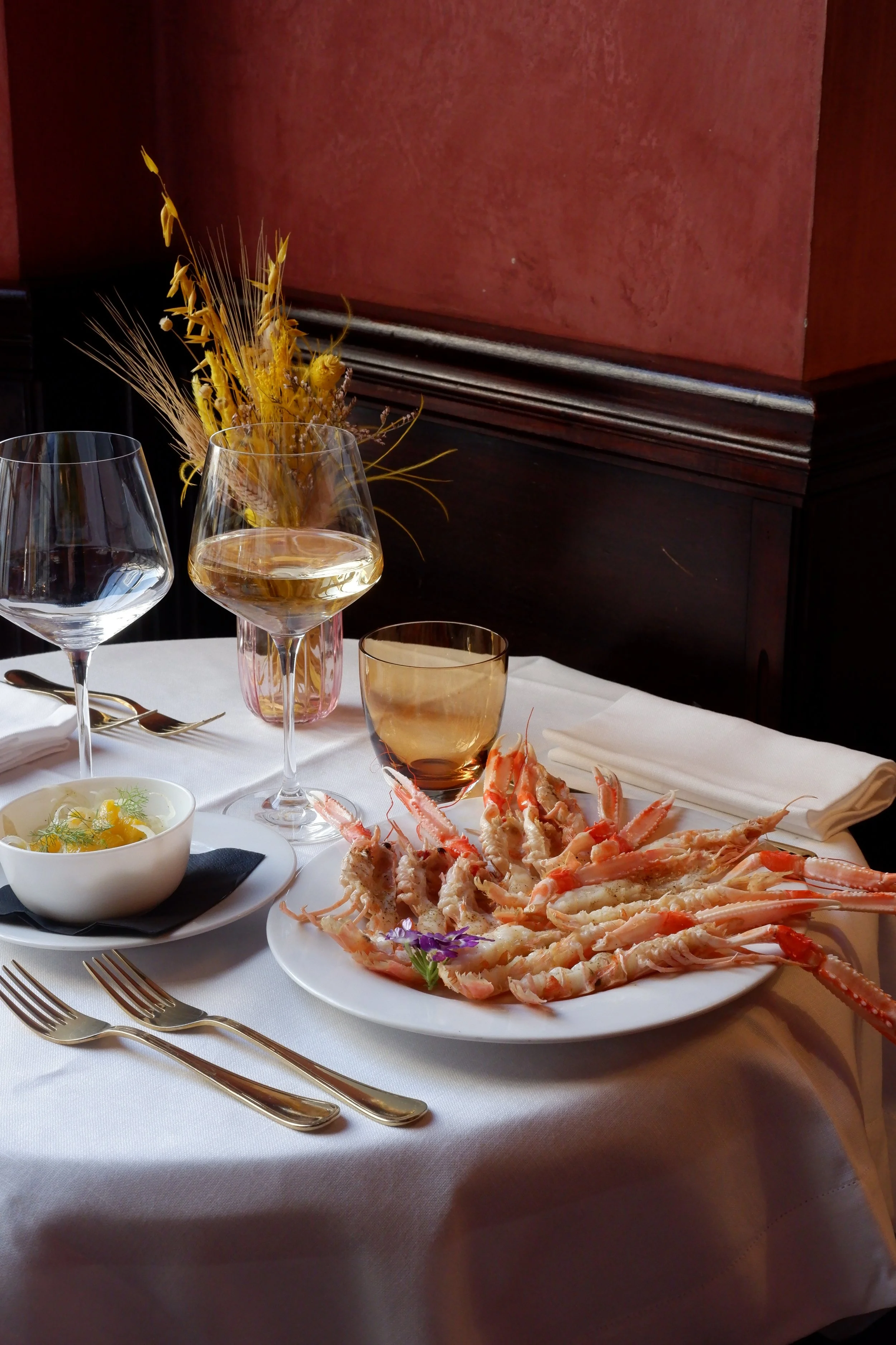 A table set with a plate of cooked lobster, a bowl of lemon and dill sauce, two wine glasses, a water glass, a drinking glass, a cup, utensils, and a flower arrangement in a pink vase against a red wall with dark wood paneling.