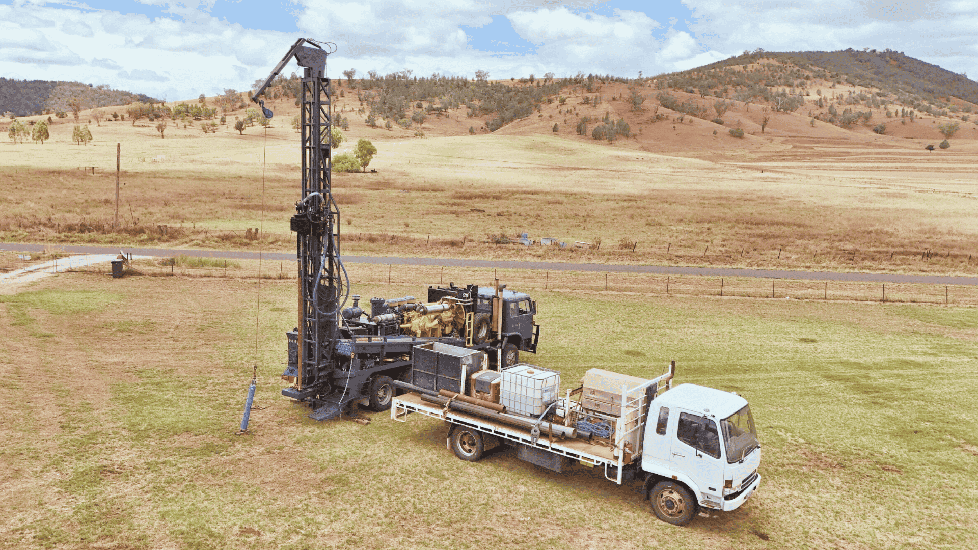 A drilling rig mounted on a truck in a rural field with hills and sparse trees in the background.