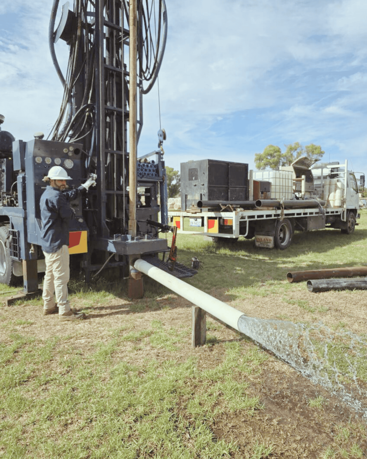 A worker in a hard hat and safety gear operates a large geothermal drilling rig, with pipes and equipment in a grassy outdoor setting under a partly cloudy sky.