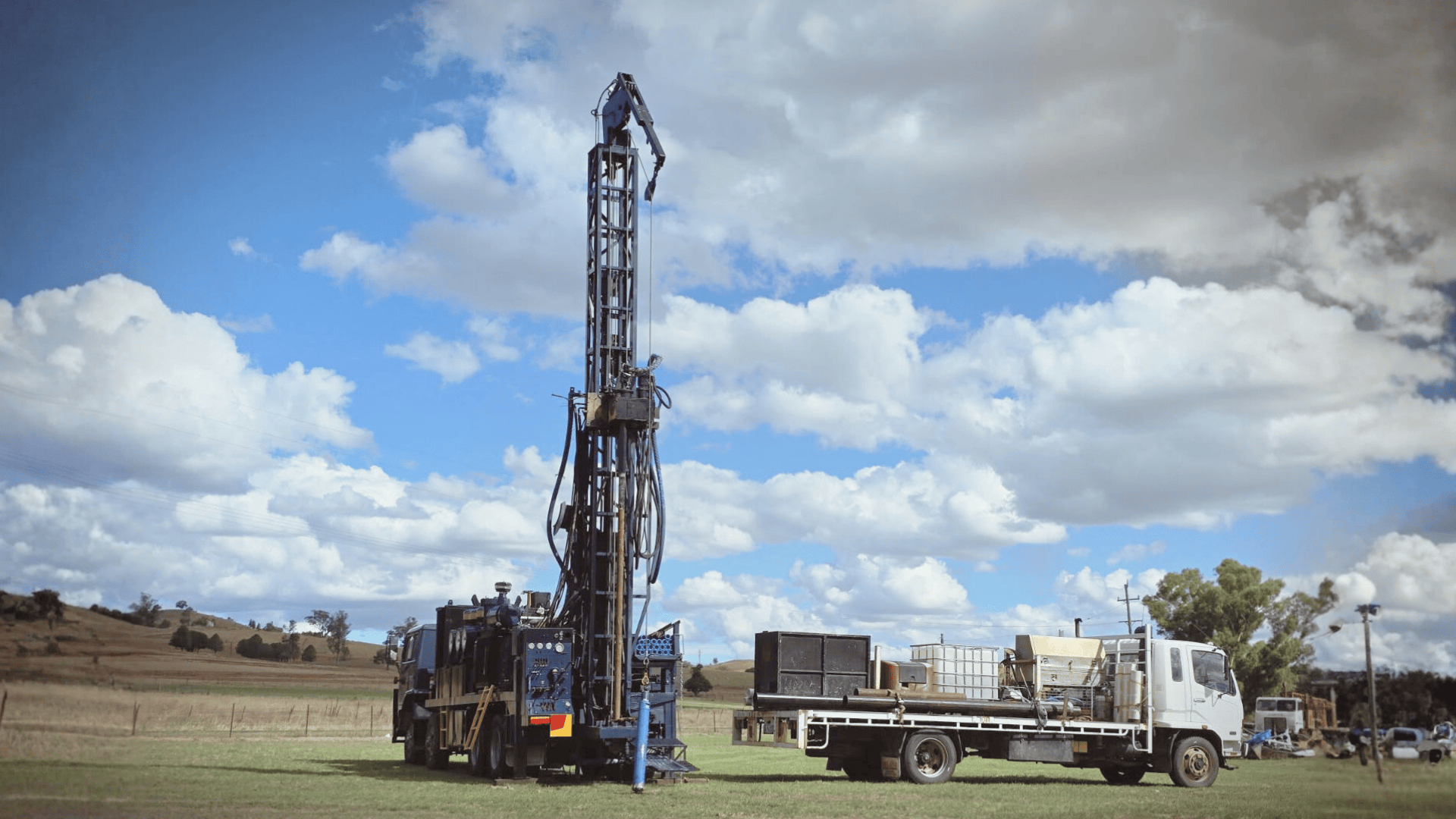 A drilling rig mounted on a truck parked in a rural field with rolling hills and scattered trees under a partly cloudy sky.