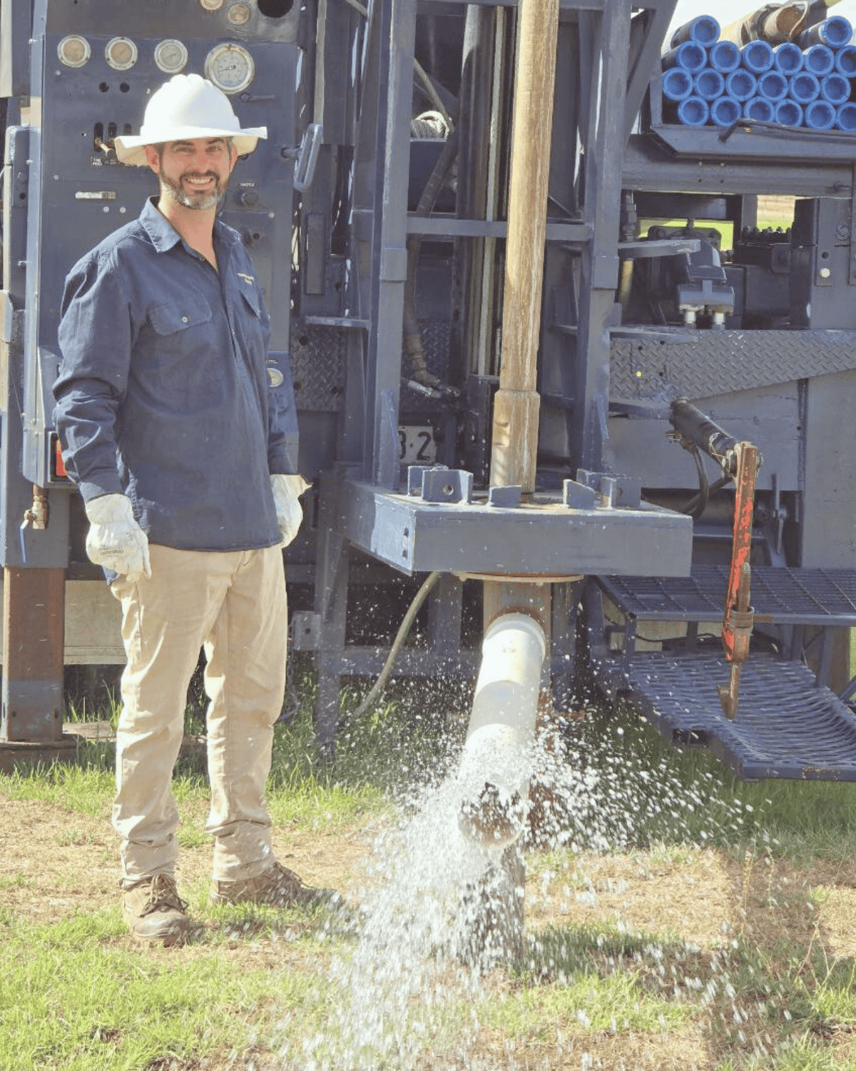 A worker in safety gear, including a white hard hat and gloves, stands next to a water well drilling rig, with water spraying from a pipe into the ground.