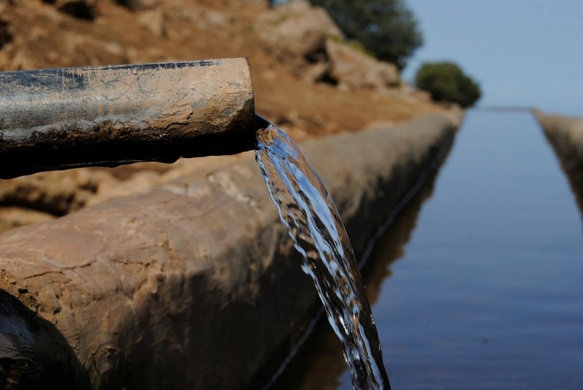Water flowing from a pipe into a canal or waterway, with a rural outdoor landscape in the background.