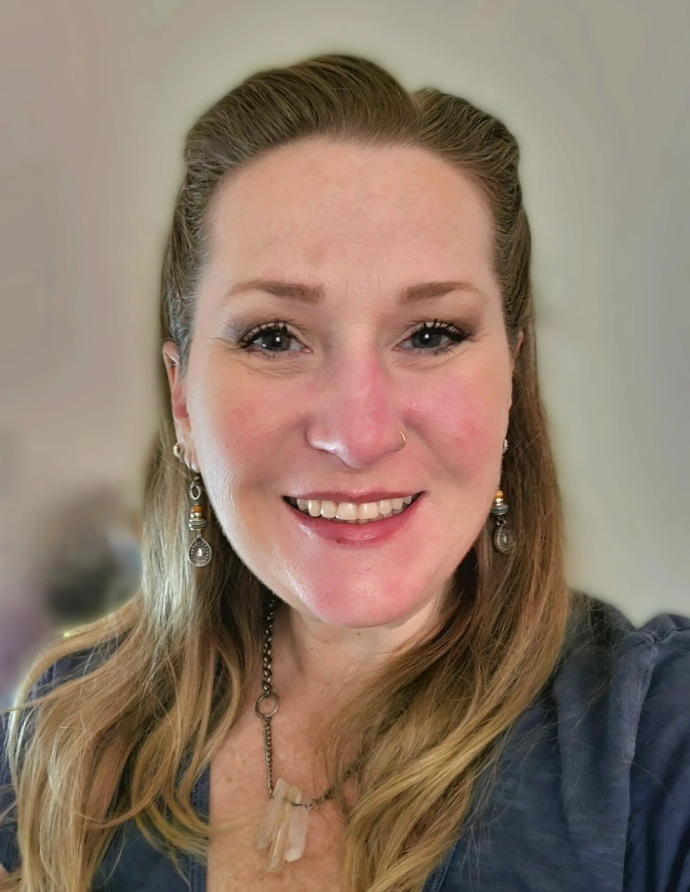 Close-up of a smiling woman with long, wavy blonde hair, wearing earrings, a necklace, and a dark top.