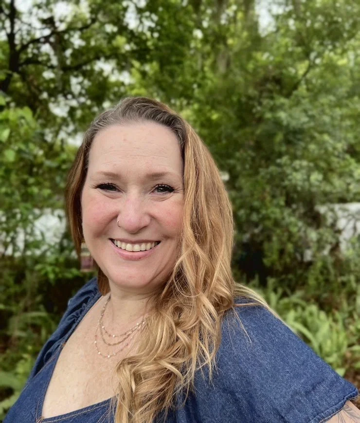 A woman with long, wavy blonde hair smiling outdoors, wearing a blue top and a layered necklace, with green trees and foliage in the background.