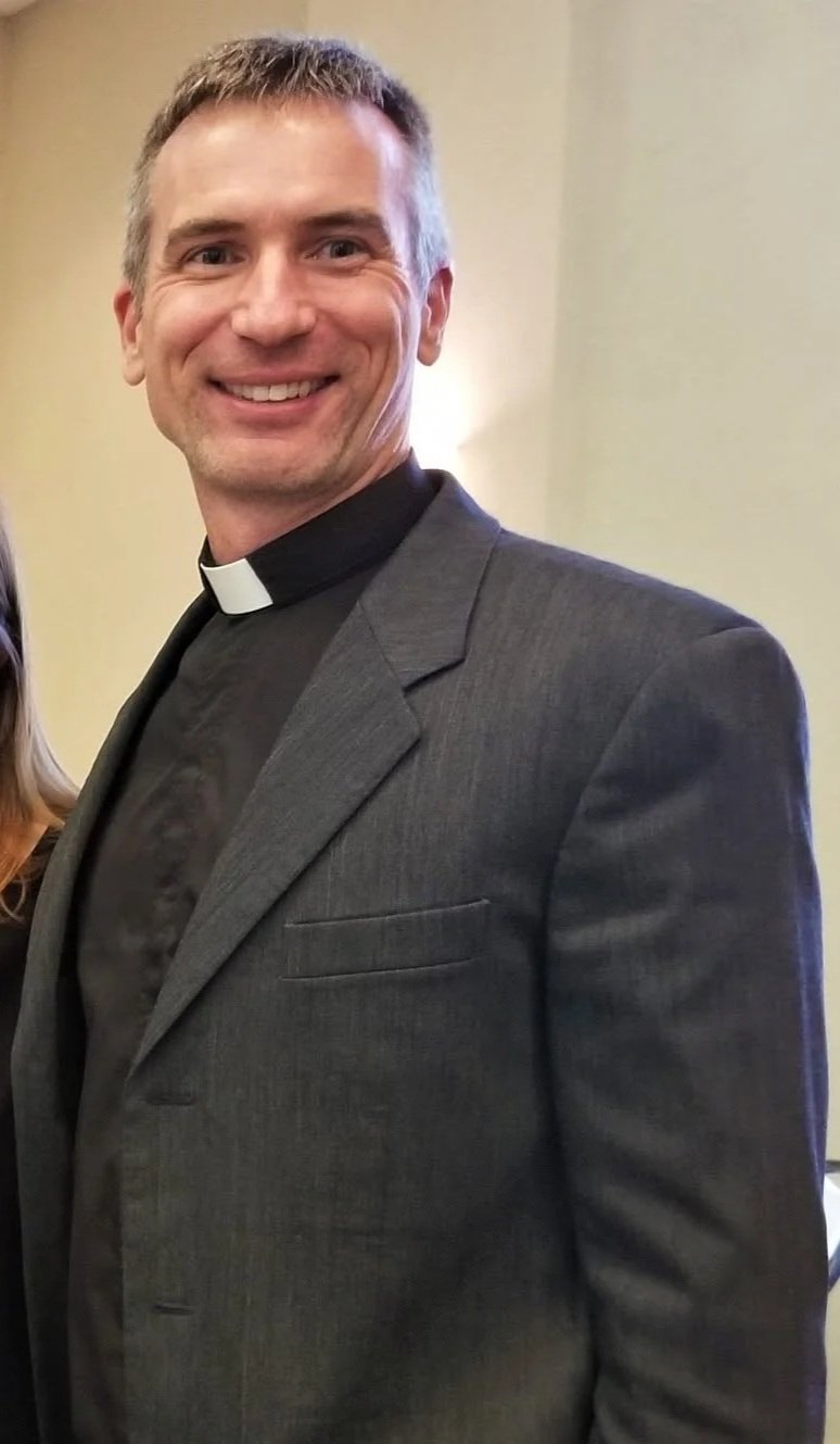 A smiling man dressed as a priest, wearing a black clerical shirt with a white collar, and a gray suit.