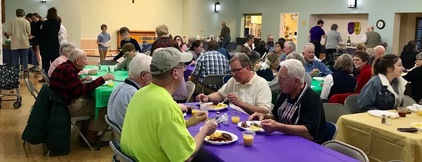 People dining at a large gathering with colorful tables and food, with many elderly and middle-aged individuals, some standing and serving food in the background.
