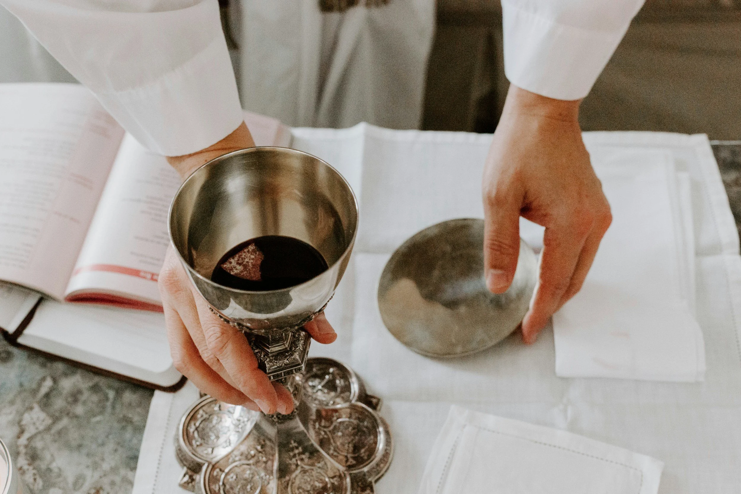 Person holding a silver goblet filled with red wine, with a book and more wine glasses on a table nearby.