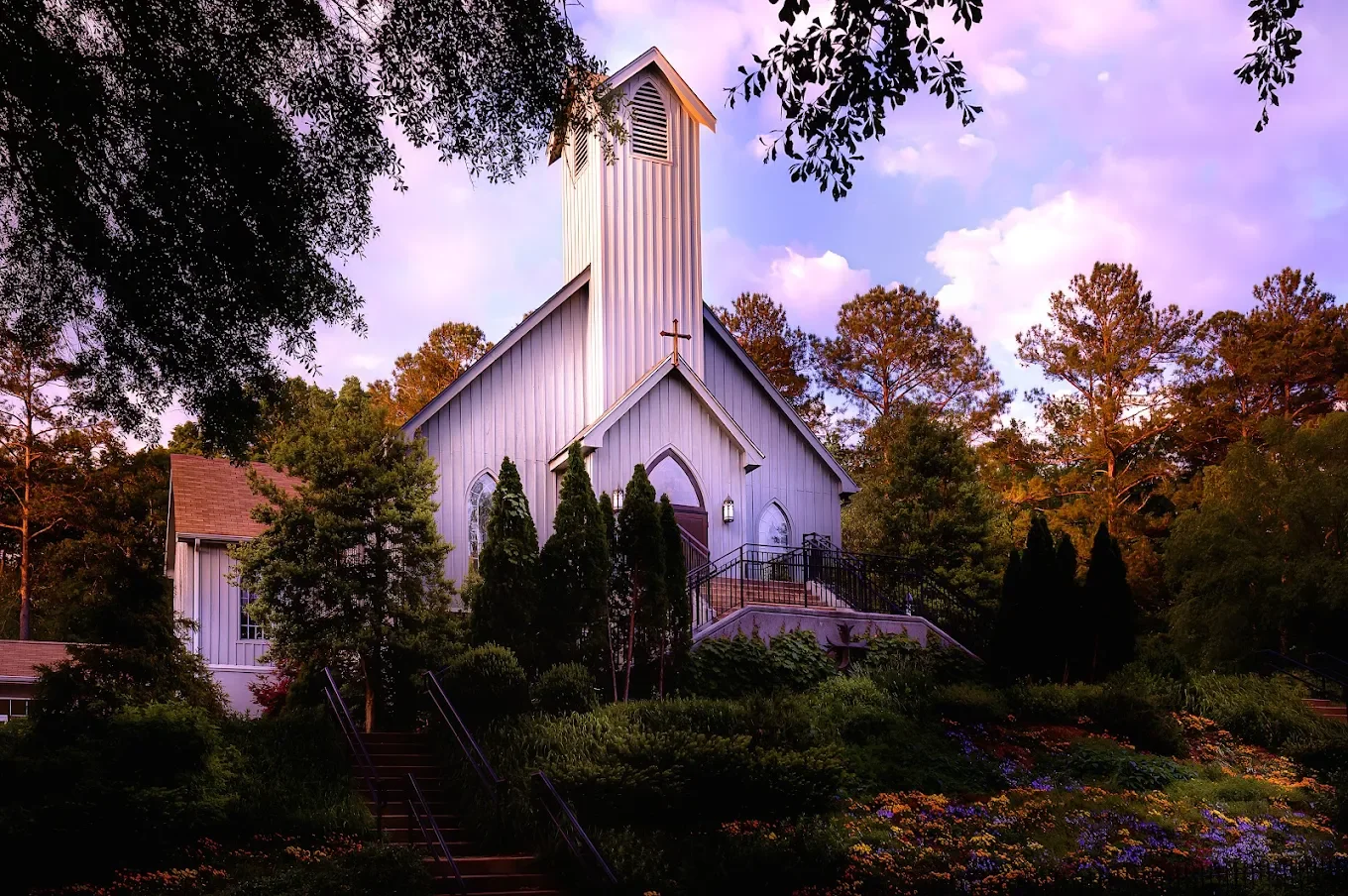 A white church with a tall steeple surrounded by trees and colorful flowers, with a pathway leading to its entrance during sunset or sunrise.