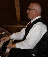 Man playing piano in a wooden room, wearing glasses, a white shirt, and a black vest.