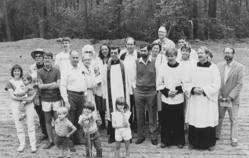 Group photo of men, women, children, and two clergy members in robes standing outdoors in a wooded area.