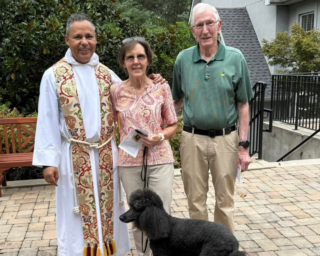 Three people and a black poodle standing outdoors on a brick patio with greenery and a building in background. One person is dressed in a religious robe, the others are casually dressed, and they are posing for the photo.