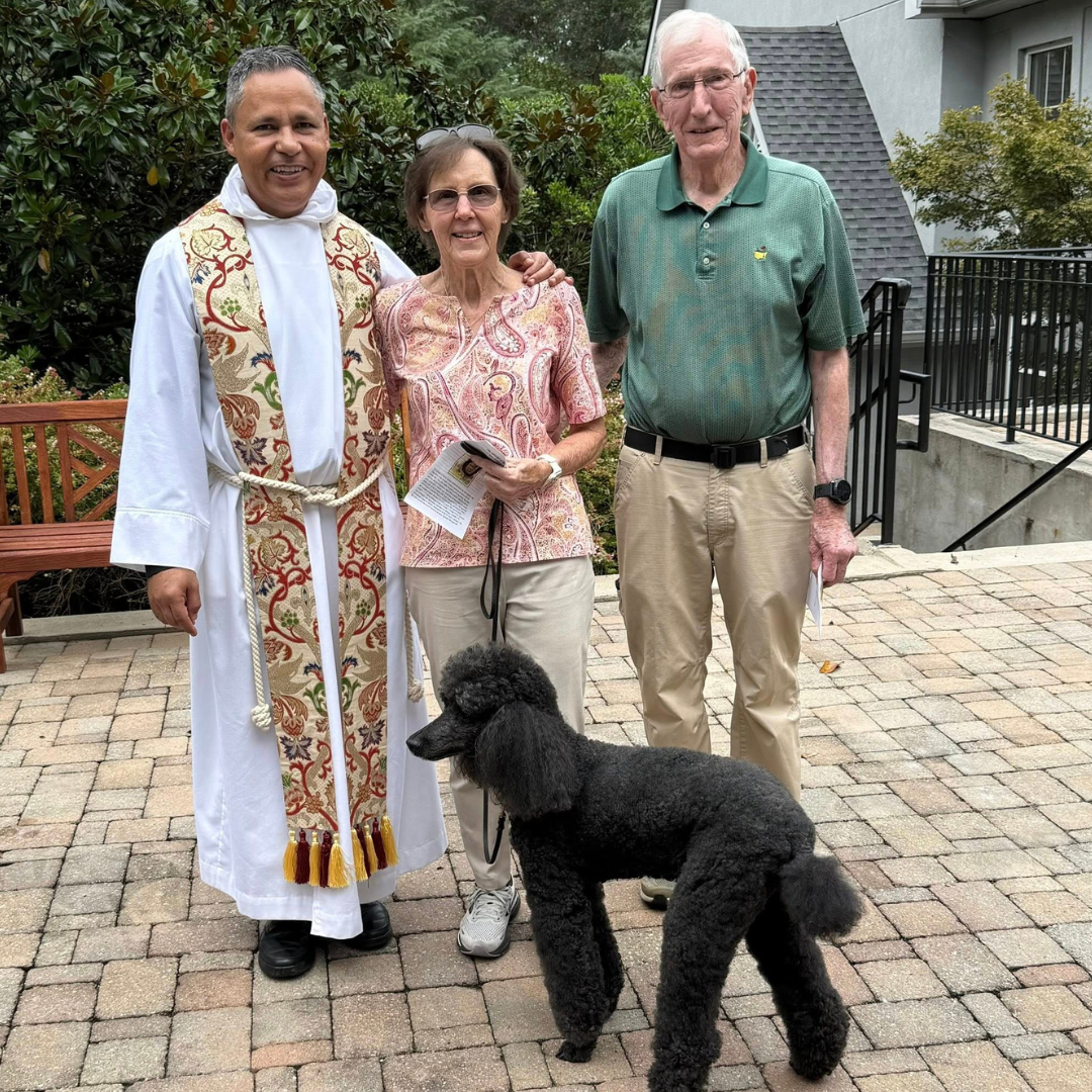 Three people standing outdoors on a brick patio, with a priest or minister on the left dressed in religious robes, a woman in the middle holding a brochure, and an older man on the right, with a black poodle dog in front of them. There are trees and a building in the background.