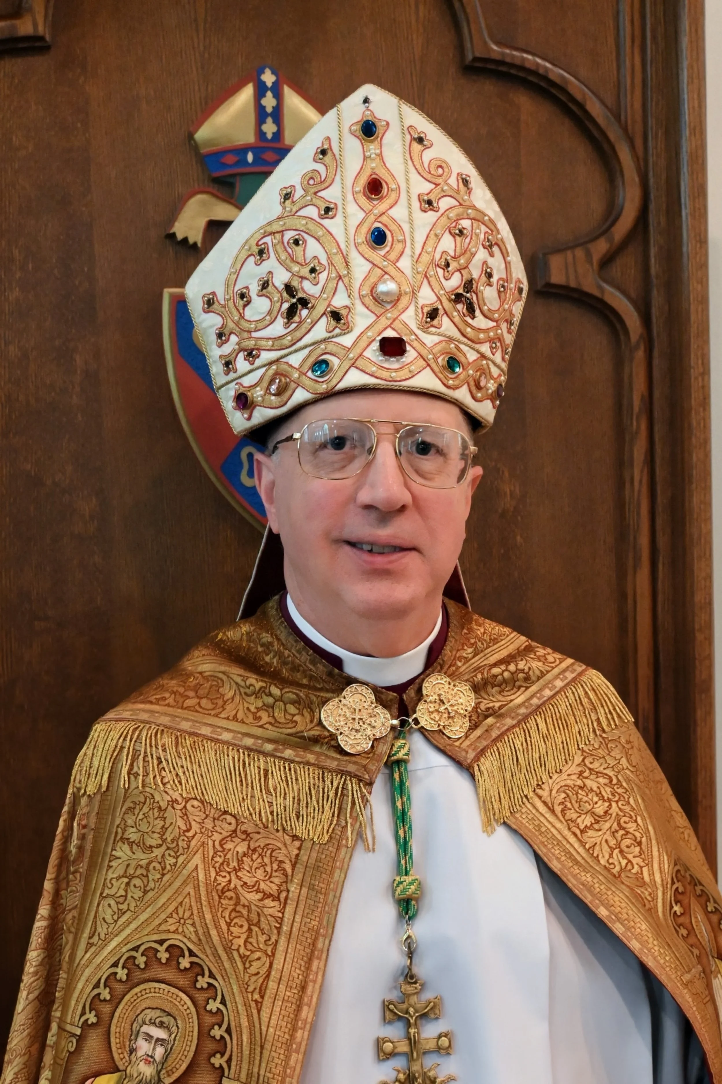 A man dressed in ornate gold and white ceremonial robes with fringes, wearing a large embroidered mitre decorated with jewels, and a long religious cross necklace. Behind him is a wooden wall with a shield emblem.