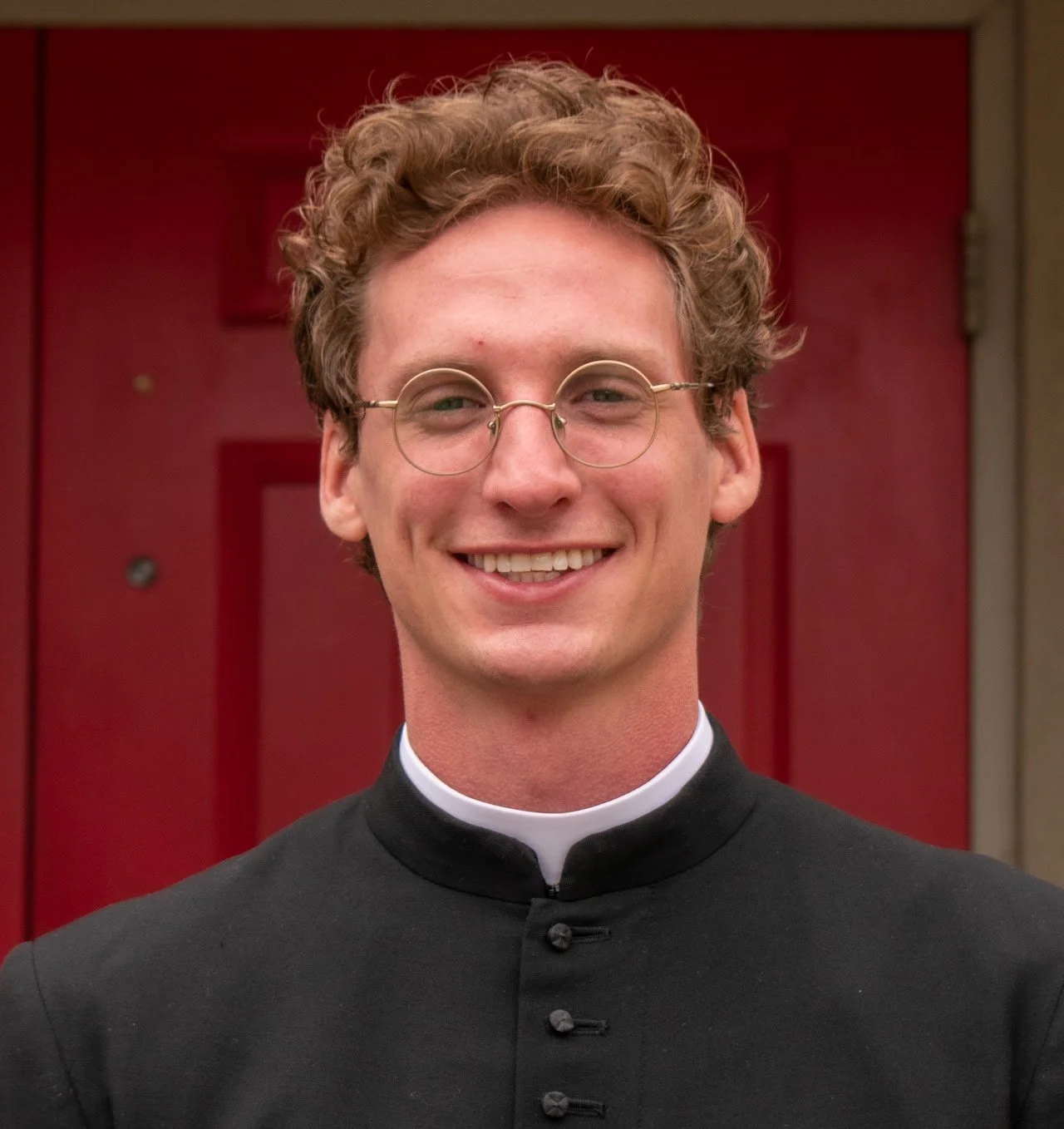 A young man with curly hair, glasses, and a clerical collar smiling in front of a red door.