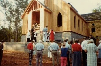 People gathered outdoors watching a construction site of a wooden church with a steeple, surrounded by trees.