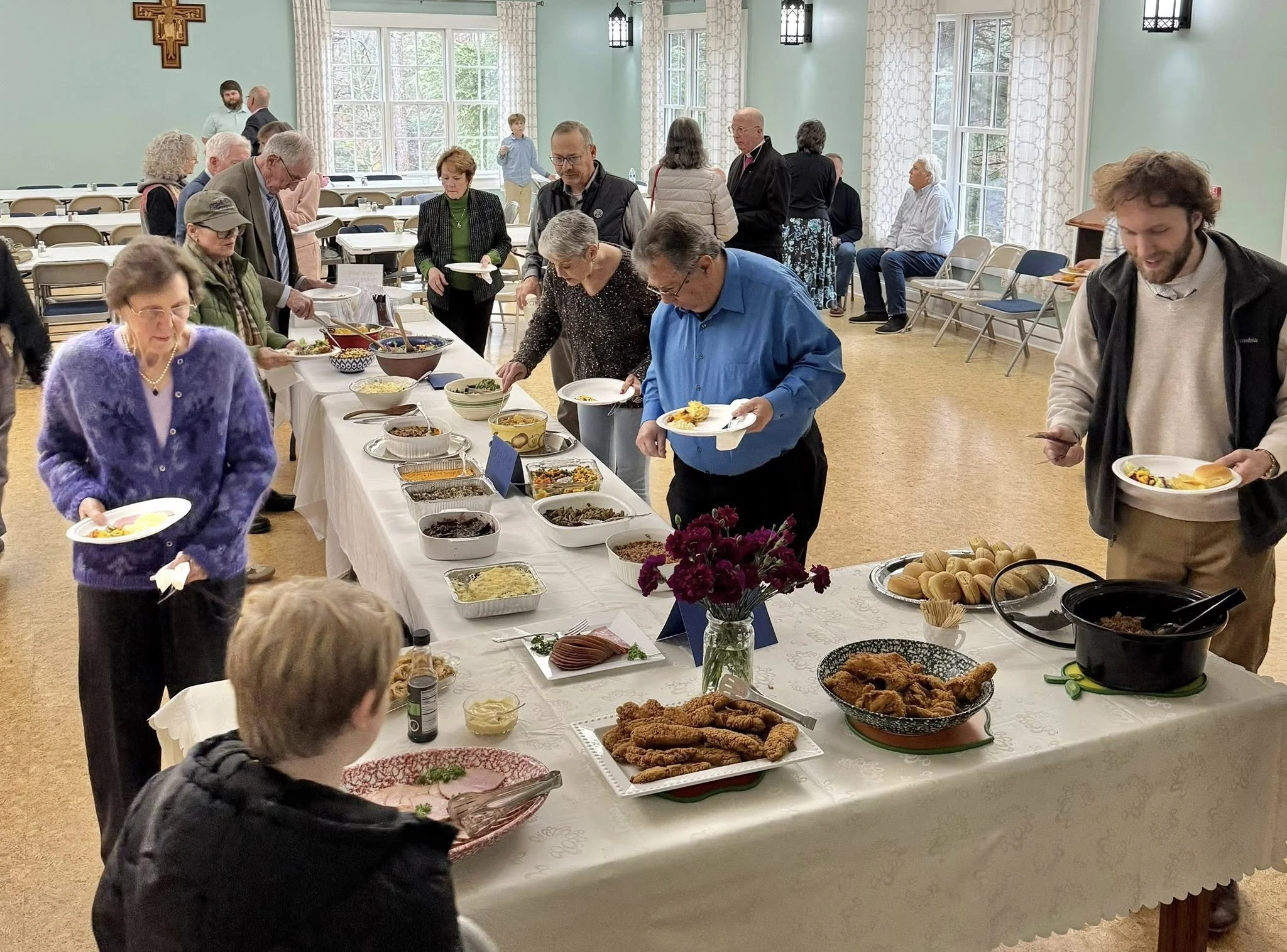 A group of people at a buffet-style meal, with various dishes including fried chicken, salads, and bread, in a room with large windows and light green walls.