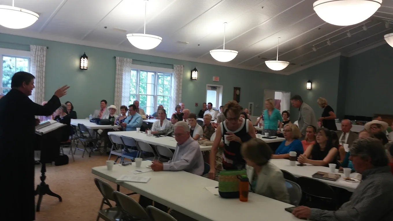 A man standing at a podium appears to be speaking to a large group of people in a conference room. The audience is seated at long tables, listening attentively. Some individuals are standing and chatting, while others are seated with drinks and notebooks in front of them. The room has light blue walls, large windows, and hanging ceiling lights.