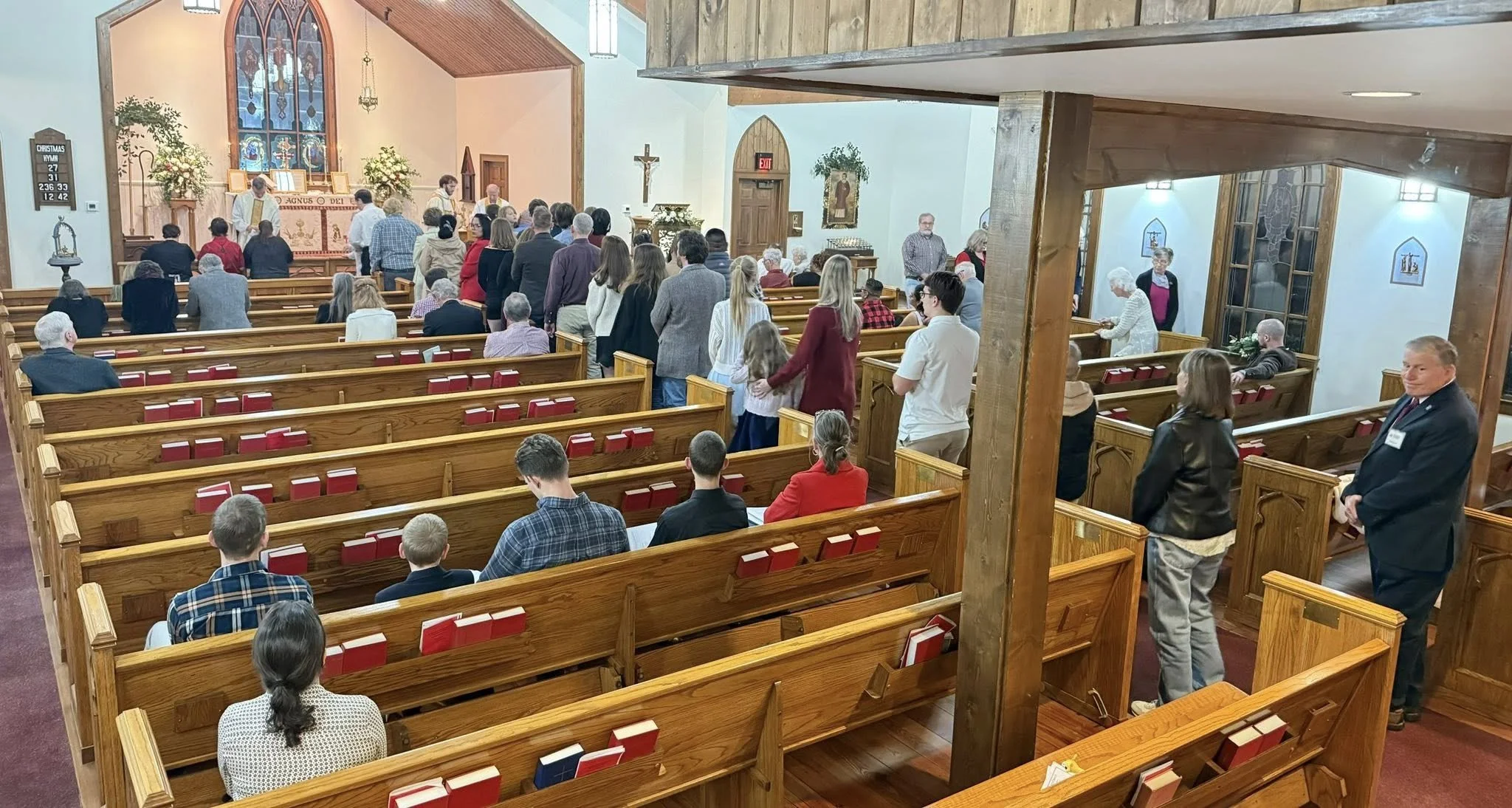 People gathered inside a church during a service, with some standing and some sitting on wooden pews. The church has stained glass windows, crucifix, and religious artwork.