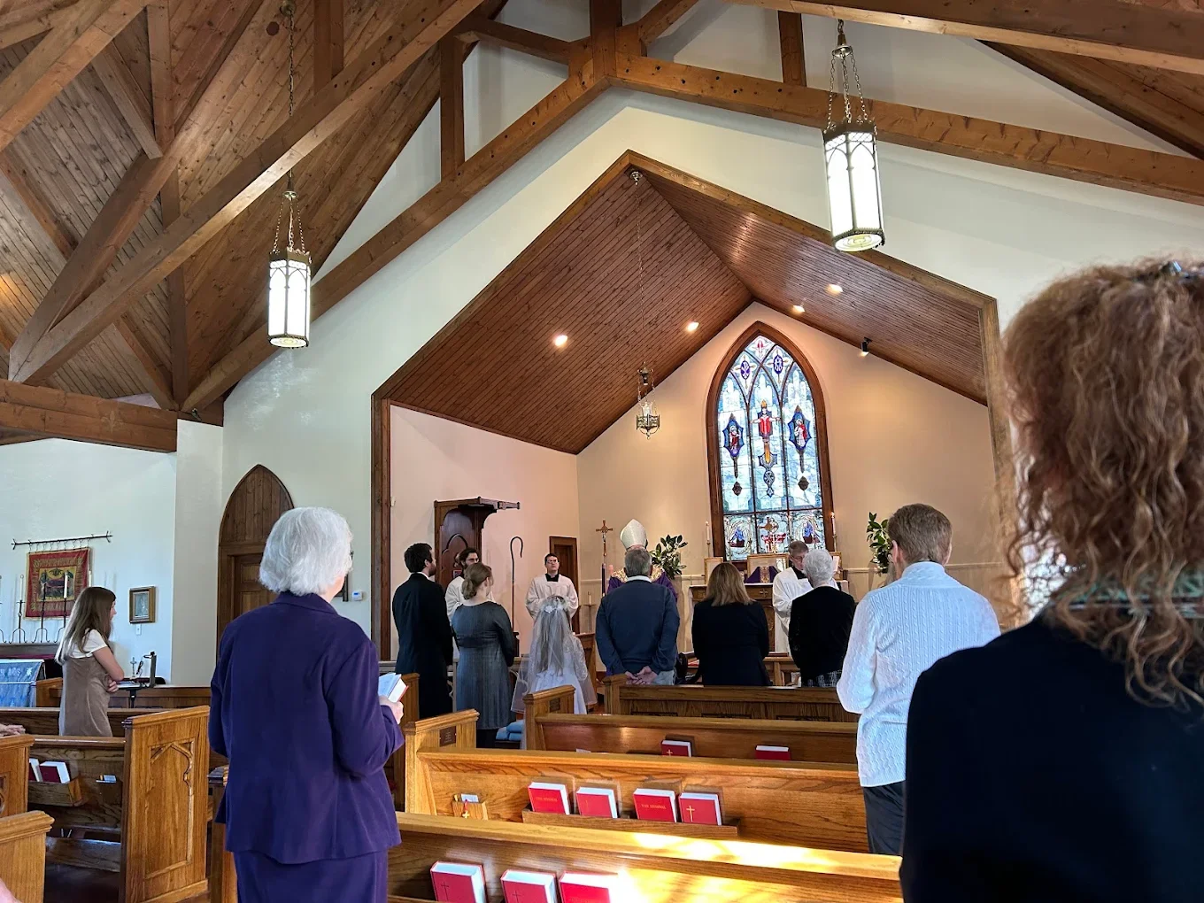 People attending a church service with an altar and stained glass window at the front, wooden pews, and a wooden ceiling with hanging lights.
