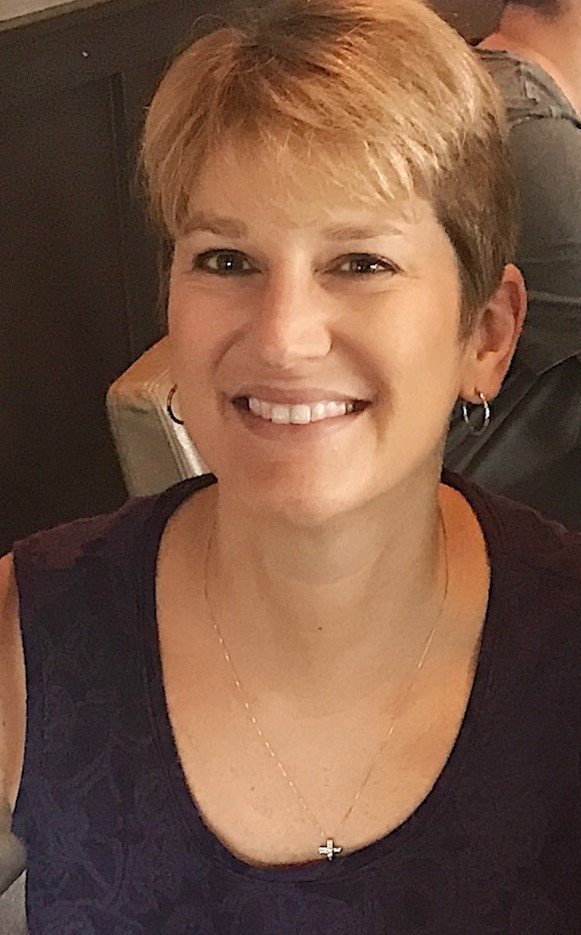 A smiling woman with short blonde hair, wearing a black top, earrings, and a silver cross necklace.