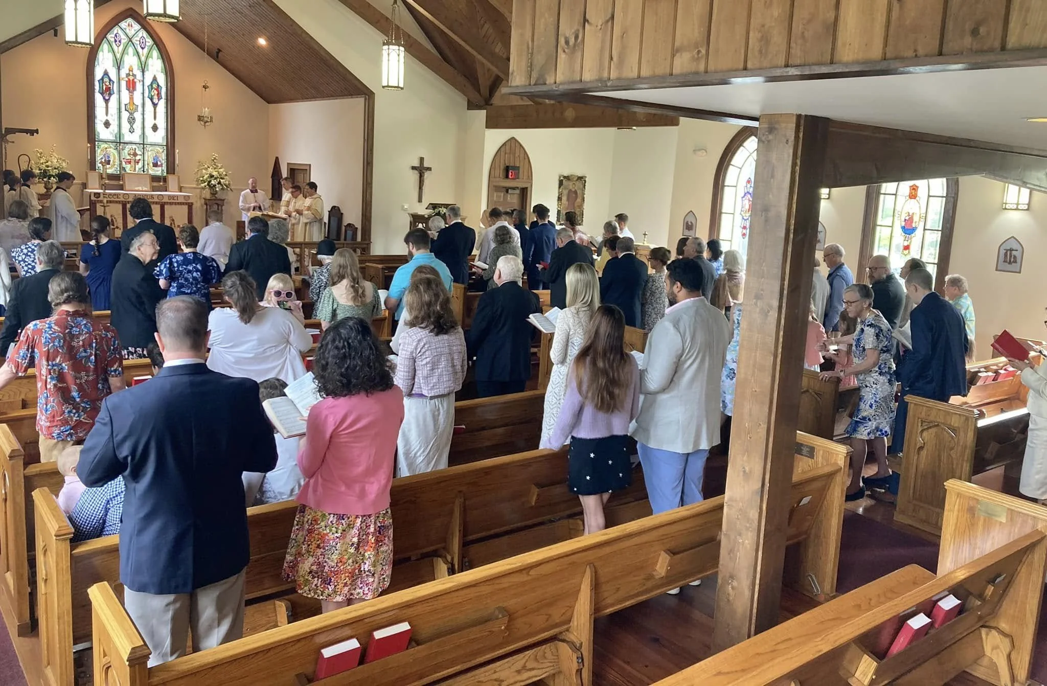 A congregation gathered inside a church during a service, with some people standing and others sitting in wooden pews. The church has stained glass windows, wooden beams, and a cross on the wall at the front. Clergy or church leaders are conducting the service at the altar.