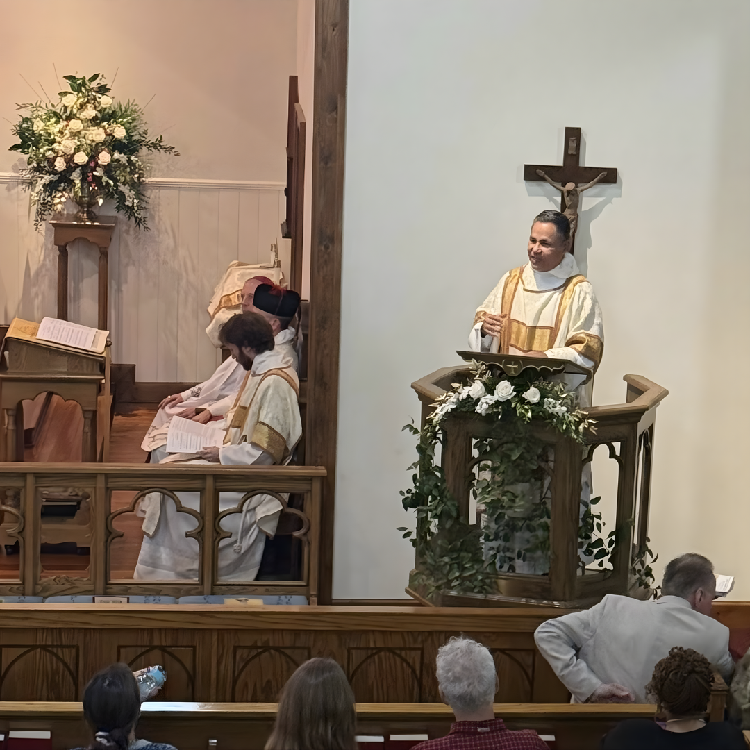 A priest delivering a sermon at a church pulpit adorned with white flowers, with a wooden cross on the wall behind him, and congregation members listening.