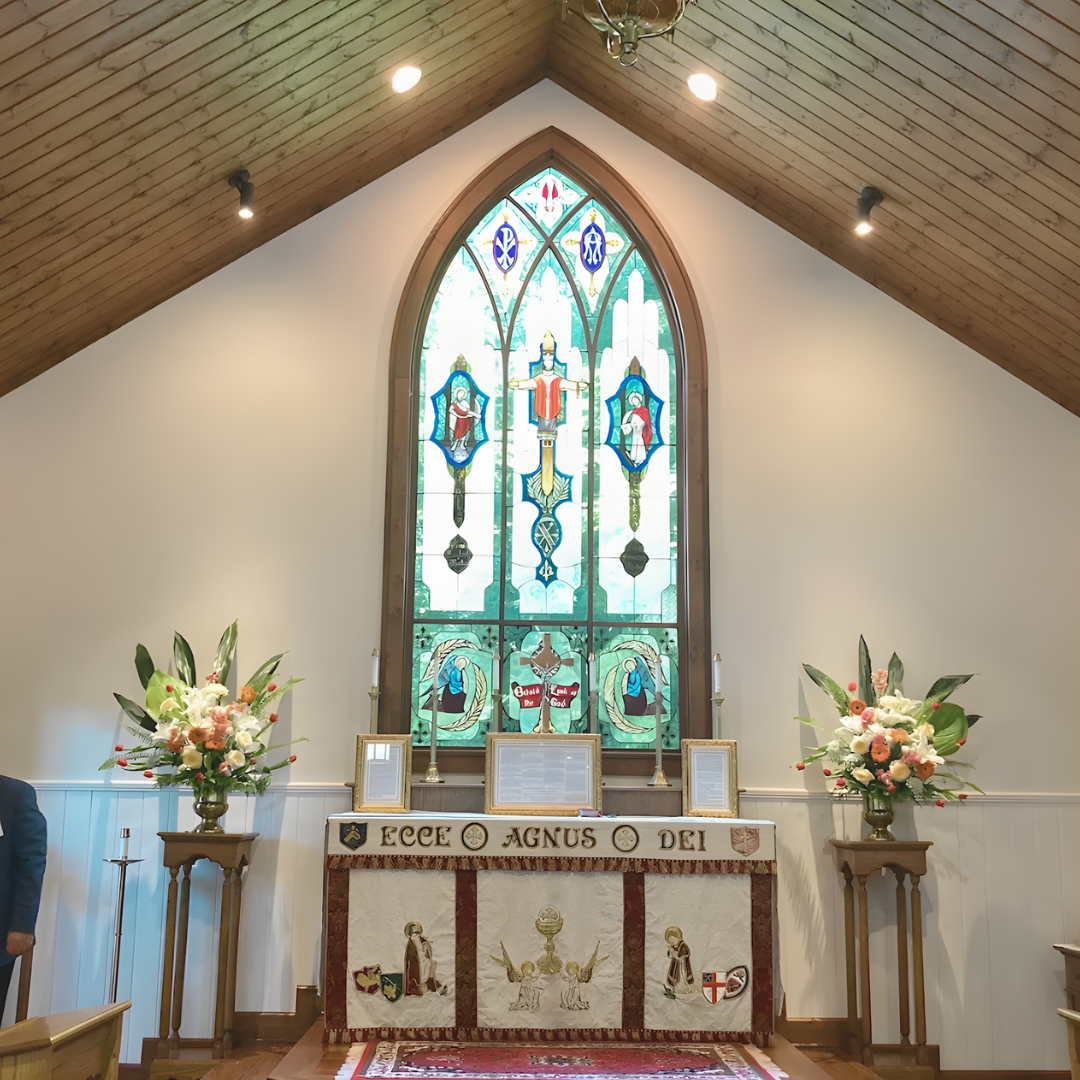 Inside a church, altar with religious banners and icons, two flower arrangements on side tables, large stained glass window depicting religious figures and symbols, wooden ceiling with spotlights.