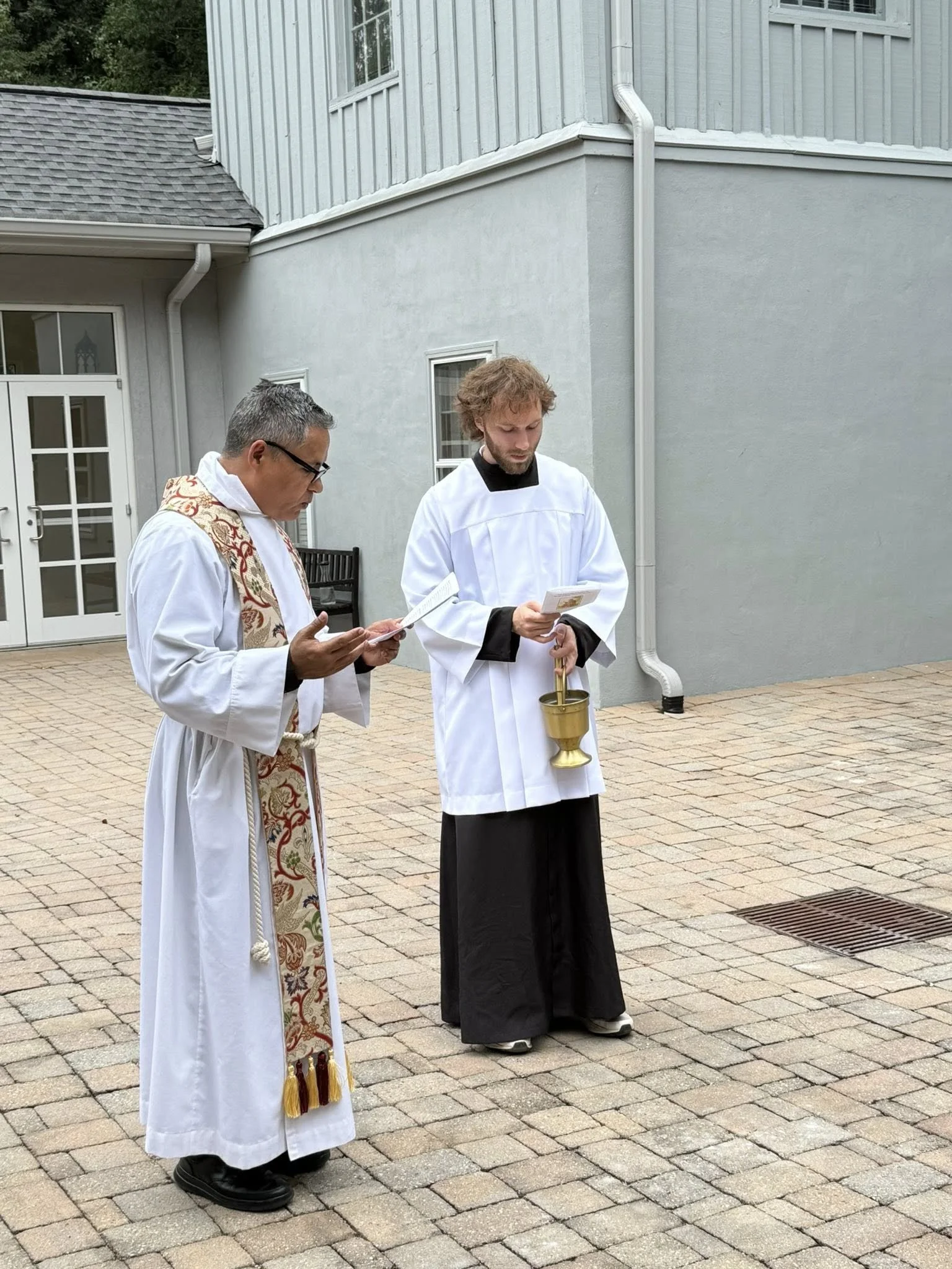 Two men dressed in religious robes stand outside on a brick patio, praying and holding papers. One man is in a white robe with a colorful stole, and the other is in a white and black cassock with a surplice. They are engaged in a prayer ritual.
