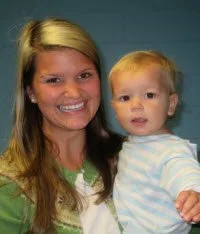 A smiling woman holding a young child with blonde hair, against a plain background.