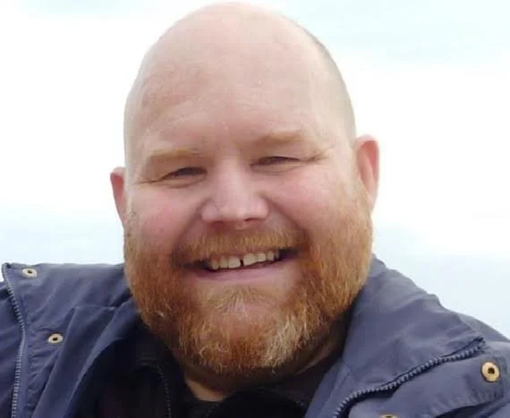 A smiling man with a beard and a shaved head, wearing a dark jacket, outdoors with a cloudy sky in the background.