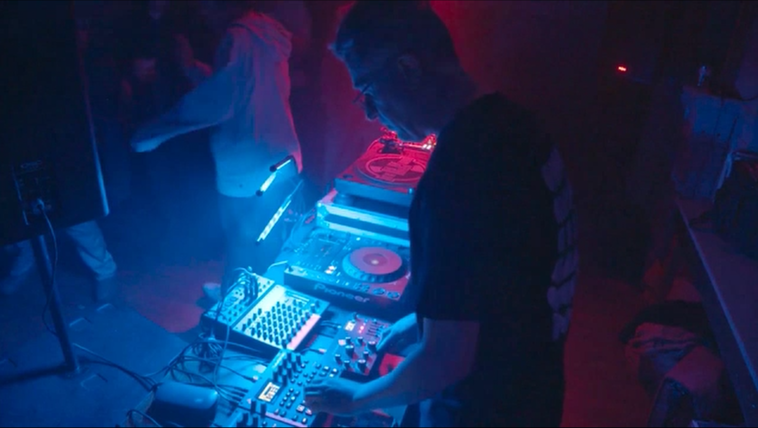 A DJ dressed in black performing in a dark club with neon lighting, using a Pioneer DJ controller and turntable, surrounded by a few people dancing.