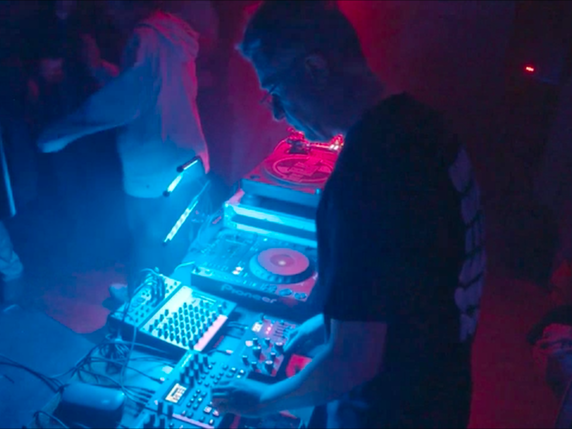 A DJ in a dark club, wearing glasses, mixes music using a Pioneer DJ controller with blue and red lighting.
