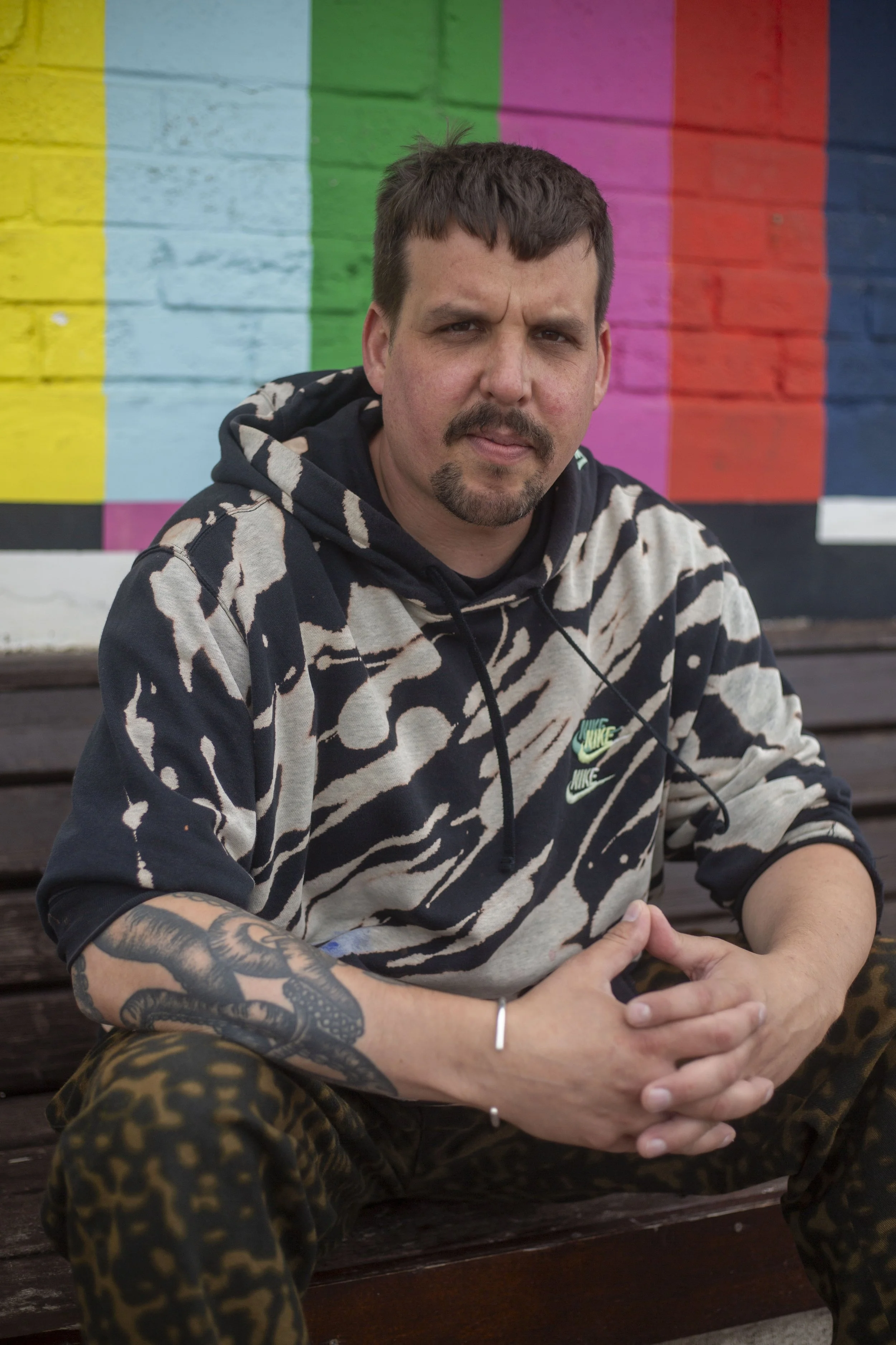 A man with short dark hair and a mustache is sitting outdoors on a bench in front of a colorful mural. He is wearing a black and beige zebra print hoodie with green Nike logos and patterned pants. His hands are clasped together, and he has a tattoo sleeve on his right arm.