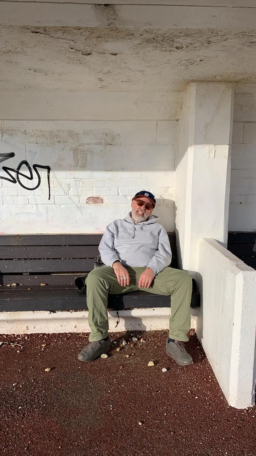 A man sitting on a bench against a white brick wall with graffiti, wearing sunglasses, a gray hoodie, khaki pants, and a black cap.
